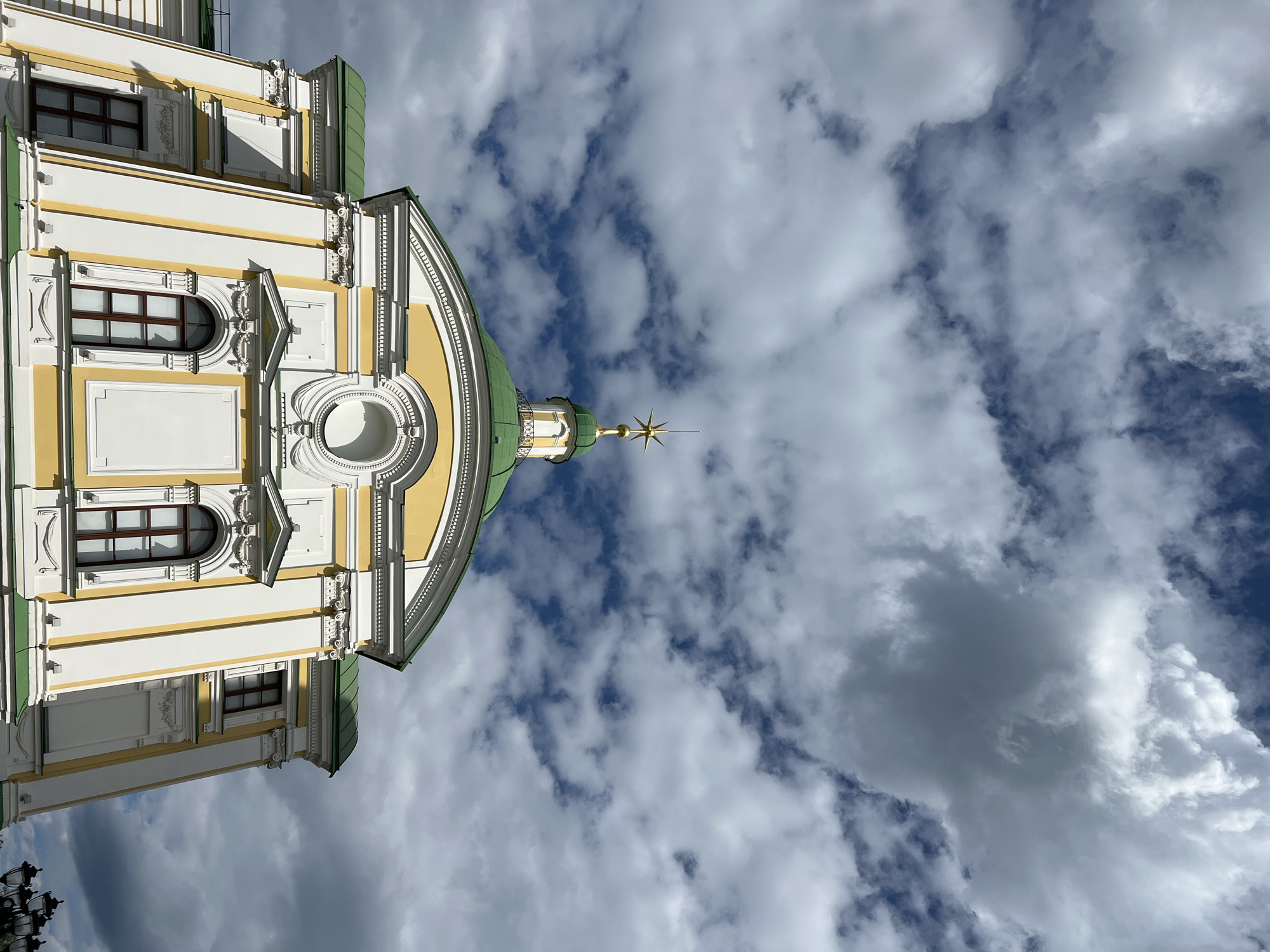 The image shows the building of the Tver Imperial Palace, a historical monument located in the city of Tver, Russia. The palace is painted white and yellow with green roofs and features intricate architectural details. A bright gold star sits atop the central green dome. The sky is filled with dramatic, fluffy clouds, suggesting a dynamic weather day. The perspective is low-angle, looking up at the palace against the sky. No people are visible in the frame. The scene is static, with the focus on the grandeur of the architecture and the atmospheric sky.