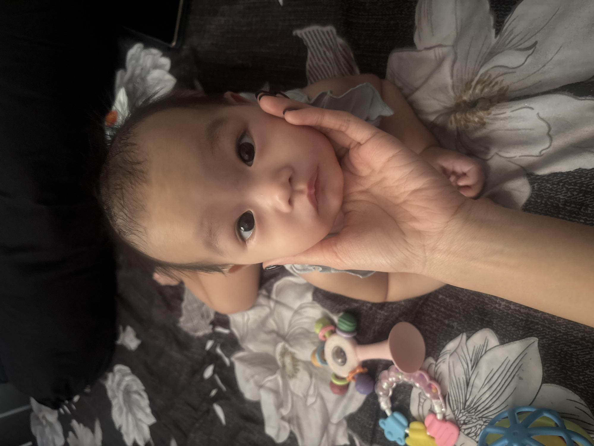 A baby is lying on a bed with a floral pattern. A person's hand is holding the baby's cheek. There are several baby toys scattered around the bed, including rattles. The baby is looking directly at the camera with big, dark eyes. The lighting in the image is dim, suggesting it might be nighttime or an indoor setting with low light. The background consists of a dark pillow and bedding. There is no specific information available in the image to determine the location in West Jakarta, Indonesia.