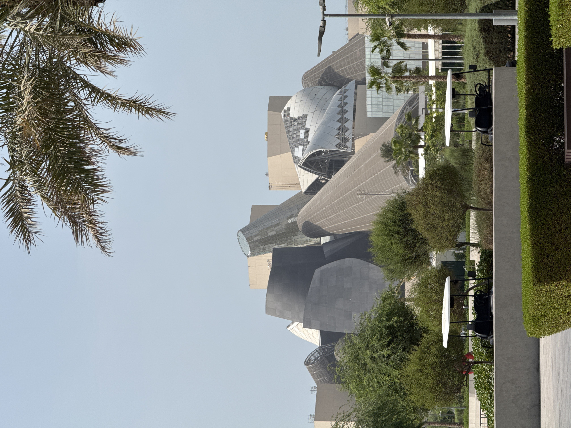 The image shows the striking architecture of the Louvre Abu Dhabi, located on Saadiyat Island in Abu Dhabi, United Arab Emirates. The museum's unique dome-like structure and interconnected buildings are visible against a clear blue sky. In the foreground, lush green trees and manicured hedges frame a concrete pathway. Two white-canopied golf carts are parked in the middle ground, suggesting a visitor or staff transport system. Palm trees and other vegetation are scattered throughout the scene, typical of the arid climate of the region. The overall impression is one of a modern, well-maintained cultural institution set within a landscaped environment.