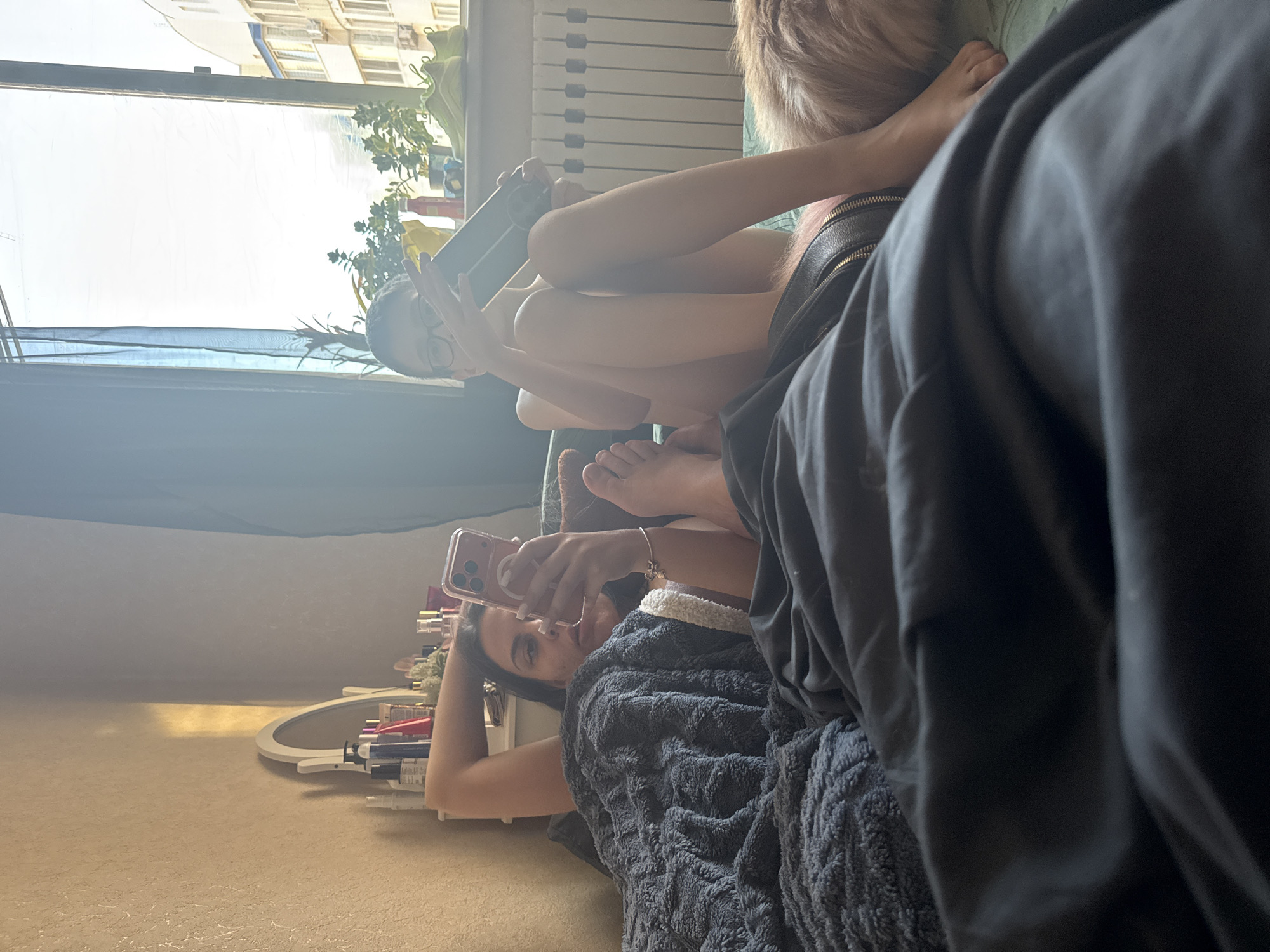 The media file shows a group of people, likely young women, relaxing indoors. One subject is lying down, covered by a textured grey blanket, and is holding a pink smartphone to take a selfie. Her left arm is resting on her head, and she is wearing a bracelet. Another subject, partially visible, appears to be sitting or lying next to her, with her legs crossed and holding a black object, possibly another phone or a gaming device. Her bare legs are prominent in the frame. A third person's upper body is visible in the background, also appearing to be relaxed. The overall scene is casual and intimate.
The setting appears to be a bedroom or living room, with a window in the background showing natural light and what looks like an apartment building outside. Sunlight streams in, creating a bright atmosphere. There is a radiator visible near the window. A vanity with makeup or toiletries is also present in the lower part of the frame. The time of day seems to be daytime, given the bright natural light. The mood appears relaxed and comfortable, with the subjects engrossed in their personal devices. No visible text is present in the image.