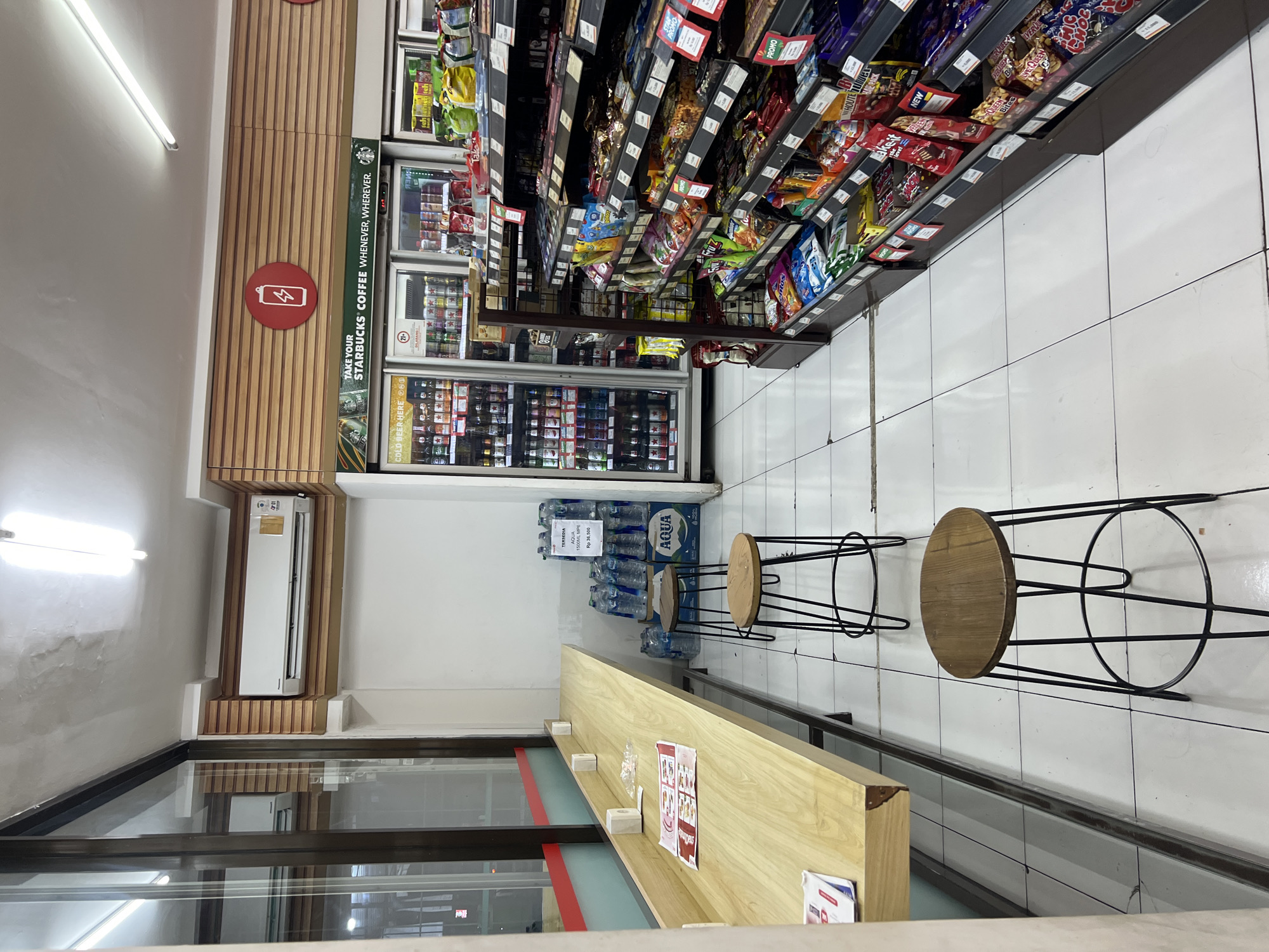The image depicts the interior of a convenience store, likely a 7-Eleven, located in Denpasar, Indonesia. The store features a long, light-wood counter with a glass partition along its length, set against a wall with horizontal wood paneling. Three tall, dark-legged stools with round wooden seats are positioned in front of the counter. To the right of the counter, there are refrigerated display cases filled with beverages, including bottles and cans. Above the refrigerated units, a sign reads "TAKE YOUR STARBUCKS COFFEE WHENEVER, WHEREVER." Adjacent to the beverage coolers are shelves stocked with various packaged snacks and confectionery items, some with "PROMO" labels. In the background, a water cooler with stacks of bottled water is visible. An air conditioning unit is mounted high on the wall above the refrigerated section. The floor is tiled with white square tiles. No individuals are present in the scene.