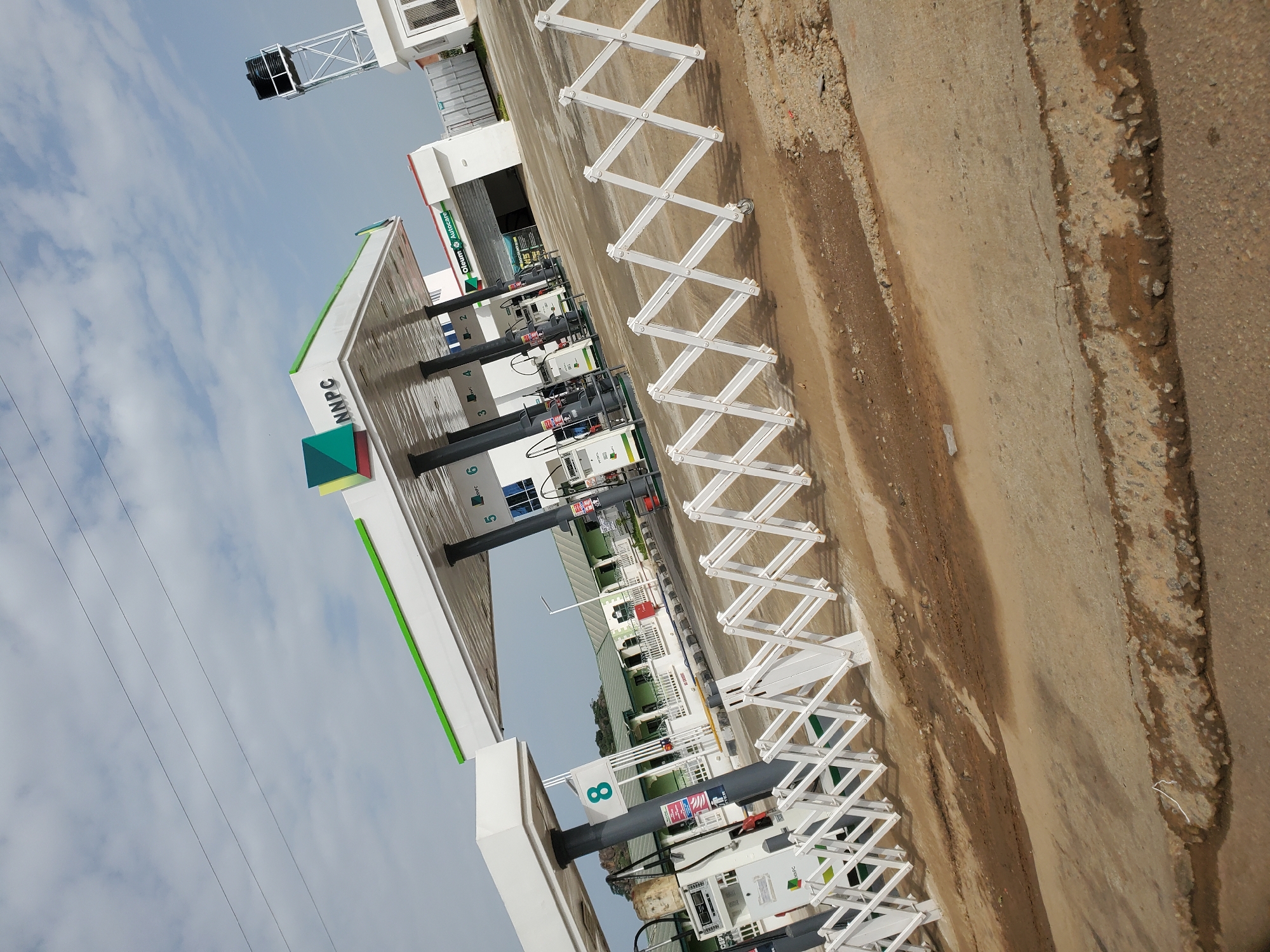 The image captures an NNPC gas station in Bukuru, Nigeria, under a partly cloudy sky. The station features a large white canopy with green trim, supported by dark gray pillars. Several gas pumps are visible under the canopy, with one clearly displaying the number "8" on its side. An expandable white metal barrier stretches across the foreground, separating the forecourt from the viewer's position. In the background, buildings with green roofs and white walls are visible, along with a tall water tower. The ground is paved with concrete and asphalt, with areas of dirt and puddles indicating recent rain or maintenance. The scene is devoid of people.