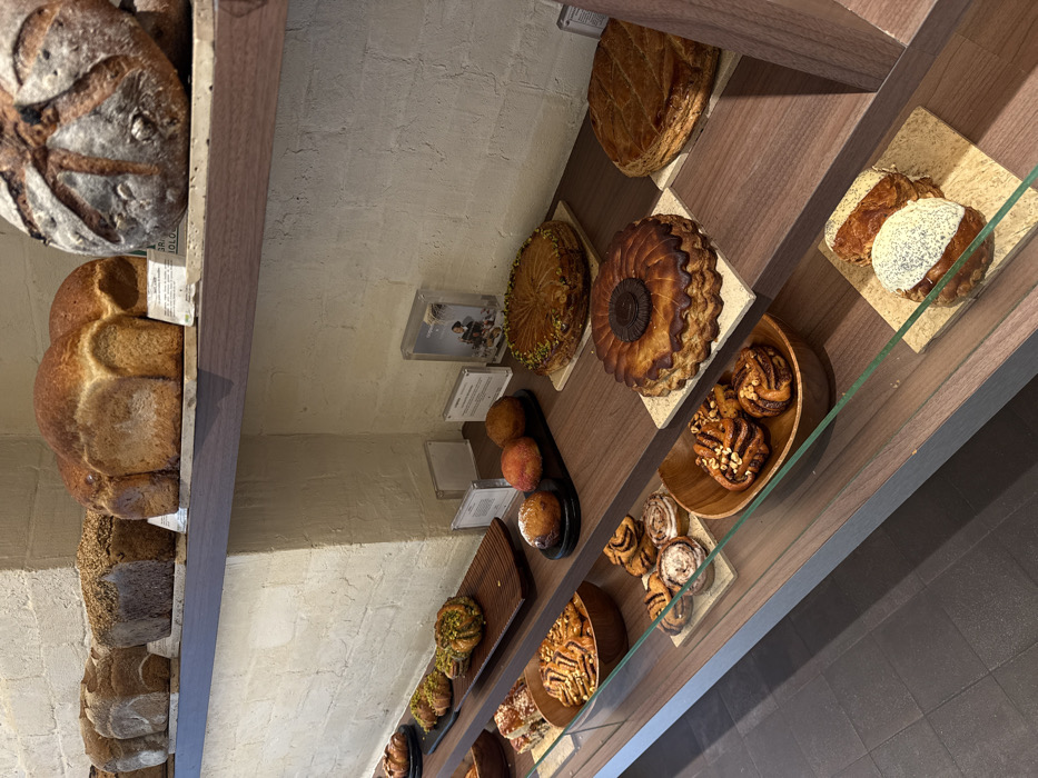 The image displays an interior view of a bakery display case within Maison Plume, located in Paris, France.
The scene features multiple shelves laden with various baked goods against a textured, light beige brick wall.
The top shelf presents several loaves of bread. From left to right: three rectangular, dark-crusted loaves, one covered in seeds; two lighter, golden-brown square loaves; and a single round, dark-crusted loaf with a scored top dusted with flour. Small white labels are positioned beneath some loaves.
The middle shelf showcases pastries and cakes. On the far left, a long, dark wooden tray holds five golden-brown pastries topped with green, likely pistachios. To the right, a black oval tray contains three round, golden-brown pastries, one appearing sugar-dusted. Further right, three large, round, golden-brown tarts or cakes are displayed on light beige square tiles. The leftmost cake has a green-dusted, intricately scored top. The central cake features a fluted edge and a dark brown circular disk in its center. The rightmost cake has a diamond-scored top. Several clear acrylic stands with white information cards are interspersed among the items; one card displays "Maison Plume" and a graphic.
The bottom shelf, partially obscured by a glass panel, holds smaller pastries. A wooden bowl on the left contains multiple swirled pastries. Adjacent to it, several smaller swirled pastries rest on a light beige tile. To the right, another wooden bowl holds multiple swirled pastries topped with chopped nuts. On the far right, two rectangular pastries are displayed on a light beige tile; the front pastry is topped with white icing and dark sprinkles.
The display unit consists of dark wooden shelves and a glass front. The floor visible at the bottom is dark grey tile. No people are visible within the frame.