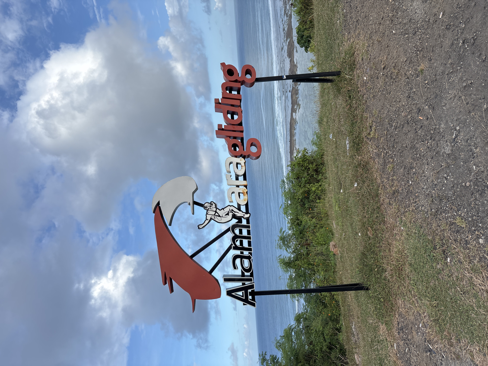 A sign for "Alam Zaa Paragliding" is set up on a grassy embankment overlooking the ocean in Kuta Selatan, Indonesia. The sign is vertically oriented with "Alam Zaa" in black letters stacked on top of each other, followed by an image of a person paragliding. Above the paraglider is a red and white wing design. Above the words "Alam Zaa" is "Paragliding" in red stacked vertically. The sign is supported by two black metal posts. The ground consists of grass, weeds, and some bare dirt. The ocean stretches to the horizon, with visible waves breaking along the shore. A strip of sandy beach is visible. Above is a cloudy sky with patches of blue.