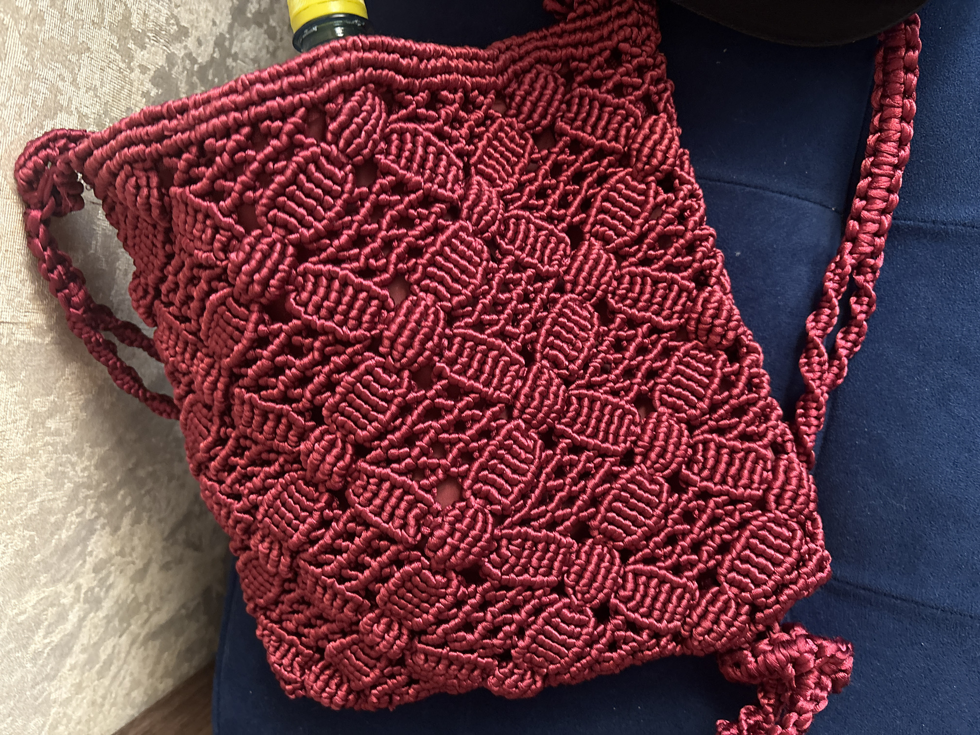 A close-up, high-angle shot features a detailed, dark red macrame bag with a patterned weave, resting on a dark blue upholstered surface. The background consists of a textured, light-colored wall. A yellow object with a black cap is partially visible on the right edge of the frame. The scene is indoors, likely in a residential setting in Georgiyevsk, Russia. No people are present in the image.