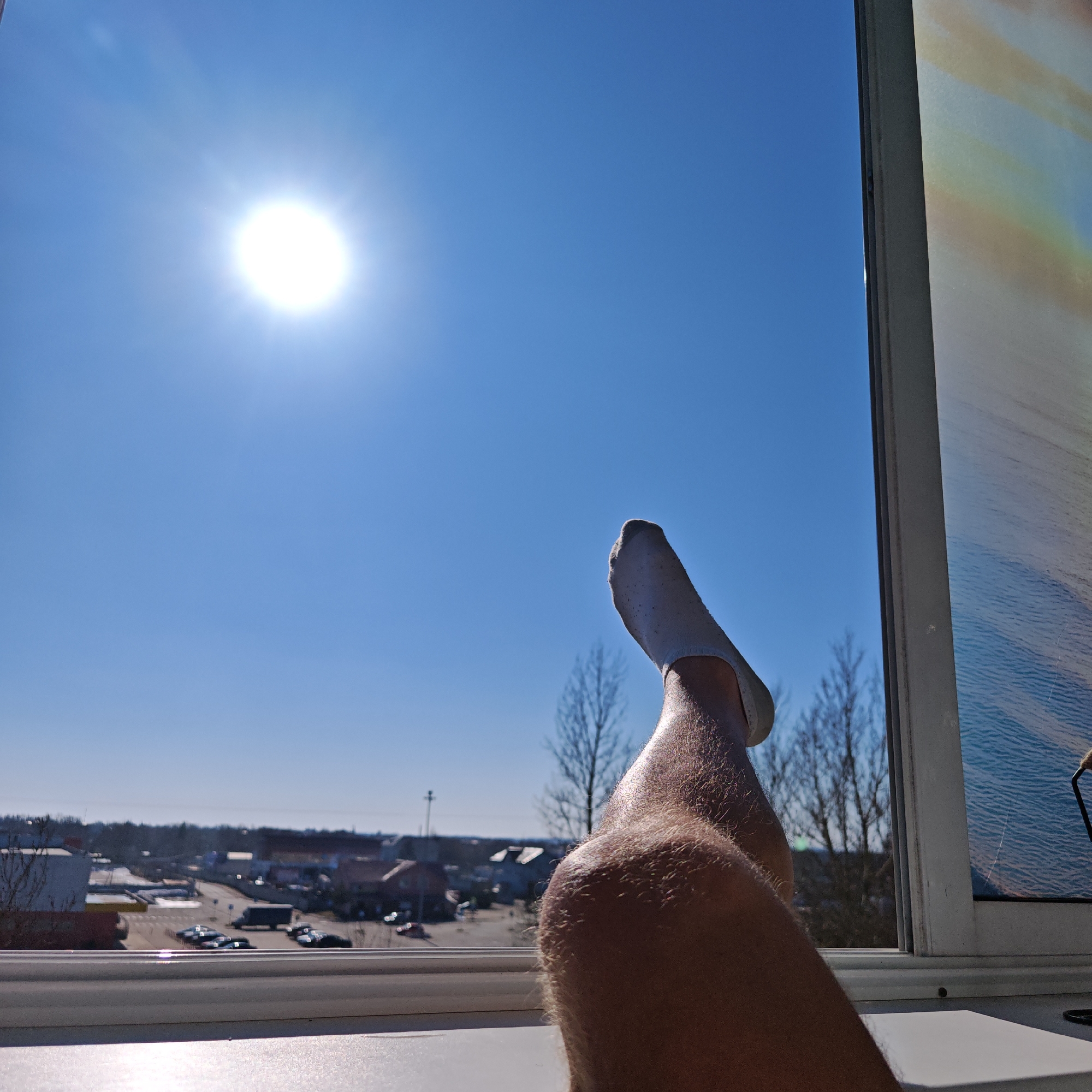 The image shows a person's leg and foot, clad in a white sock, propped up against a window frame. The view outside the window is a bright, sunny day with a clear blue sky and a distant urban landscape, including buildings, parking lots with cars, and bare trees. The bright sun is visible in the upper left of the frame. The window frame is white and has a secondary window with a picture of the sea on it. The location is not recognizable as Rostov, Russia.