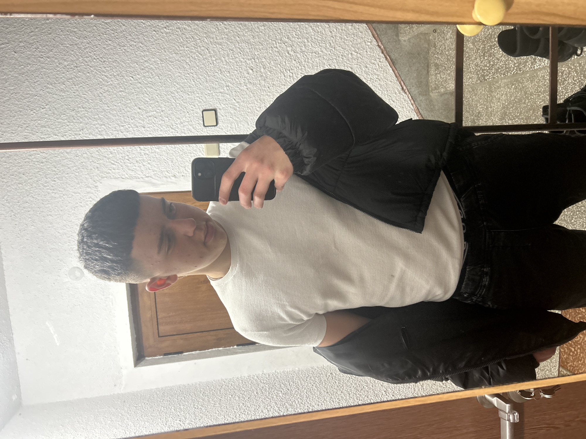 A young man stands indoors, facing a mirror, holding a black smartphone to take a selfie. He wears a white long-sleeved waffle shirt, a black puffer jacket, and black jeans. The interior appears to be a hallway or room with white walls and wooden trim. Behind him, reflected in the mirror, are stairs with a grey textured surface, and a partial view of a metal rack with clothes. The location cannot be identified as being in Zlatograd, Bulgaria from the image.