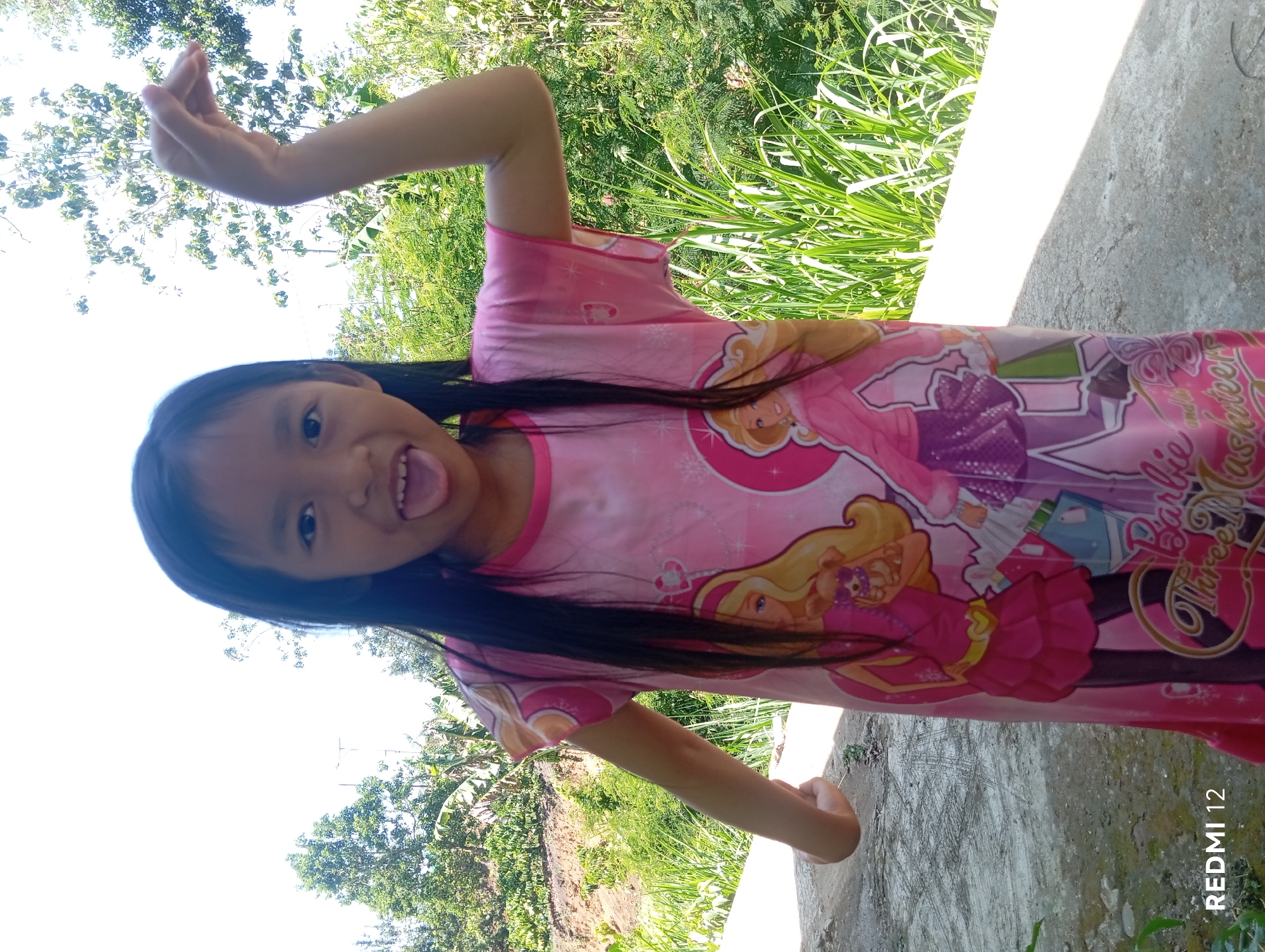 A young Asian girl, appearing to be around 5-7 years old, stands outdoors on a concrete surface. She is wearing a pink dress featuring graphics of Barbie characters. The girl has long, dark hair and is sticking her tongue out, with her right arm raised in a playful pose. Behind her, a lush green environment with trees and vegetation is visible under a bright, clear sky. The background suggests a natural, possibly rural or semi-rural setting. The bottom left corner of the image displays "REDMI 12". The scene is consistent with an outdoor location in Pagar Alam, Indonesia, known for its natural beauty and greenery.