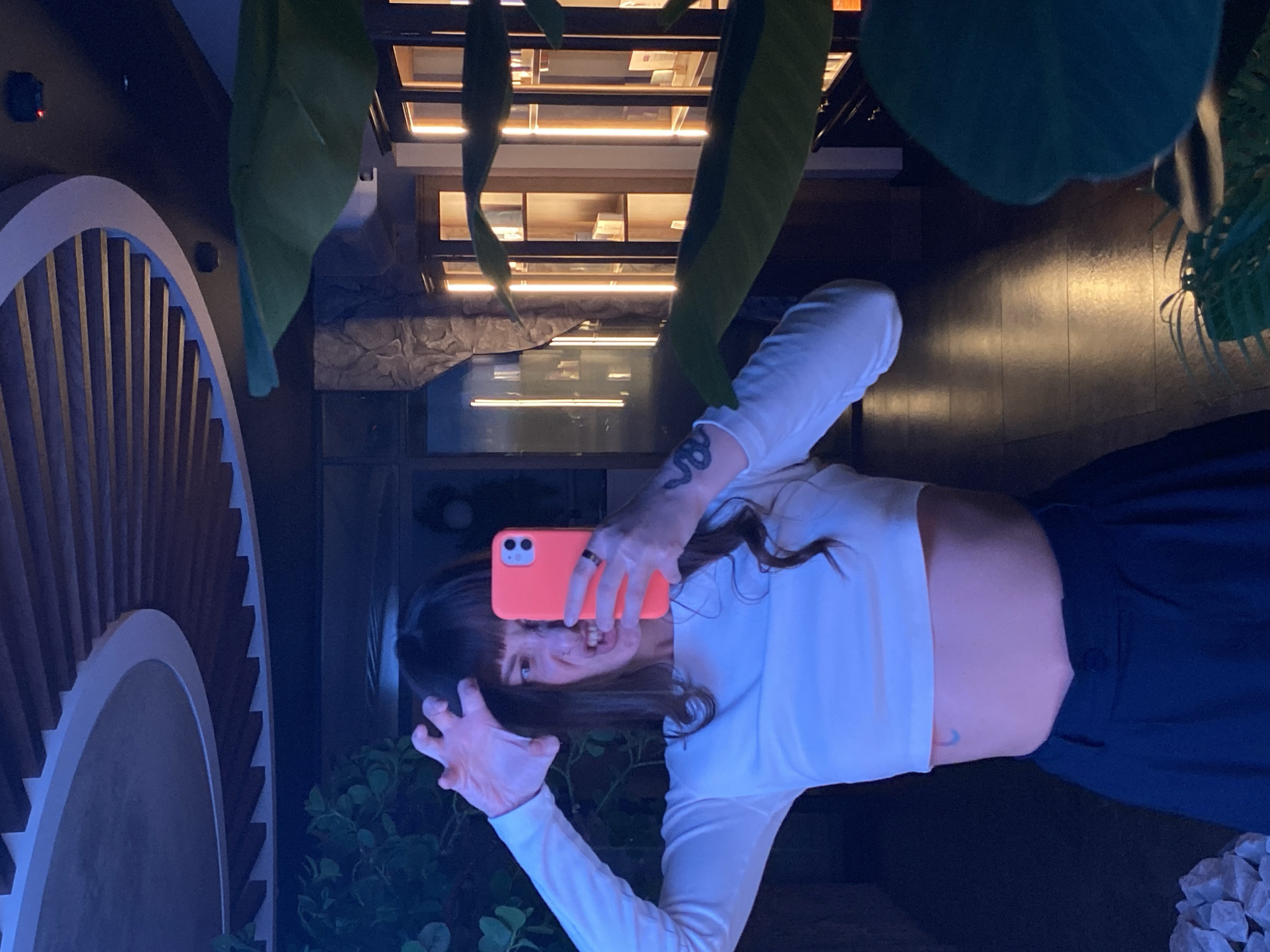 A woman is taking a selfie in a dark, possibly indoor, setting. She is holding a pink smartphone and making a claw-like gesture with one hand. She is wearing a white cropped long-sleeved shirt and dark pants. Tattoos are visible on her arm and torso. In the background, there is a decorative architectural structure resembling a saw blade, green leafy plants, a display cabinet filled with items, and dark wooden flooring. The lighting is a cool blue hue. There is no indication that this picture was taken in Votkinsk, Russia, from the content of the image.