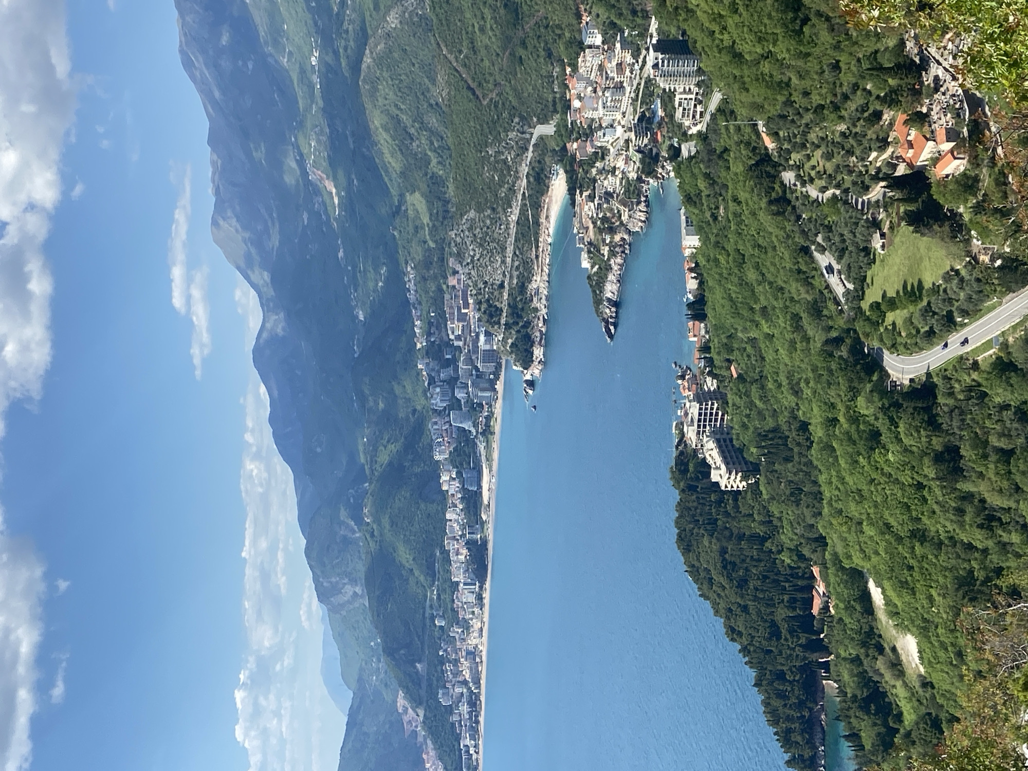 This panoramic image depicts a breathtaking coastal landscape of Sveti Stefan, Montenegro, viewed from an elevated perspective on a bright, sunny day. The deep blue Adriatic Sea spans the middle ground, its calm waters reflecting the clear sky.
In the background, majestic, rugged green mountains rise dramatically, their slopes densely covered in trees and vegetation, extending towards a light blue sky adorned with scattered white cumulus clouds. Along the left side of the coastline, a long stretch of sandy beach is visible, bordered by a line of buildings that form a town or resort.
Further to the right, the iconic islet of Sveti Stefan is prominently featured, characterized by its cluster of tightly packed, red-roofed stone buildings. It is connected to the mainland by a narrow causeway. Several boats are scattered in the turquoise waters near the coast and around the island.
The right side of the image showcases heavily forested hillsides descending towards the sea. A winding paved road snakes through these verdant slopes, with occasional buildings and structures, some with red roofs, nestled among the trees. The scene conveys a sense of serene natural beauty and a picturesque Mediterranean atmosphere, likely during morning or early afternoon. No specific human activity is distinctly visible, and no text is discernible within the image.
