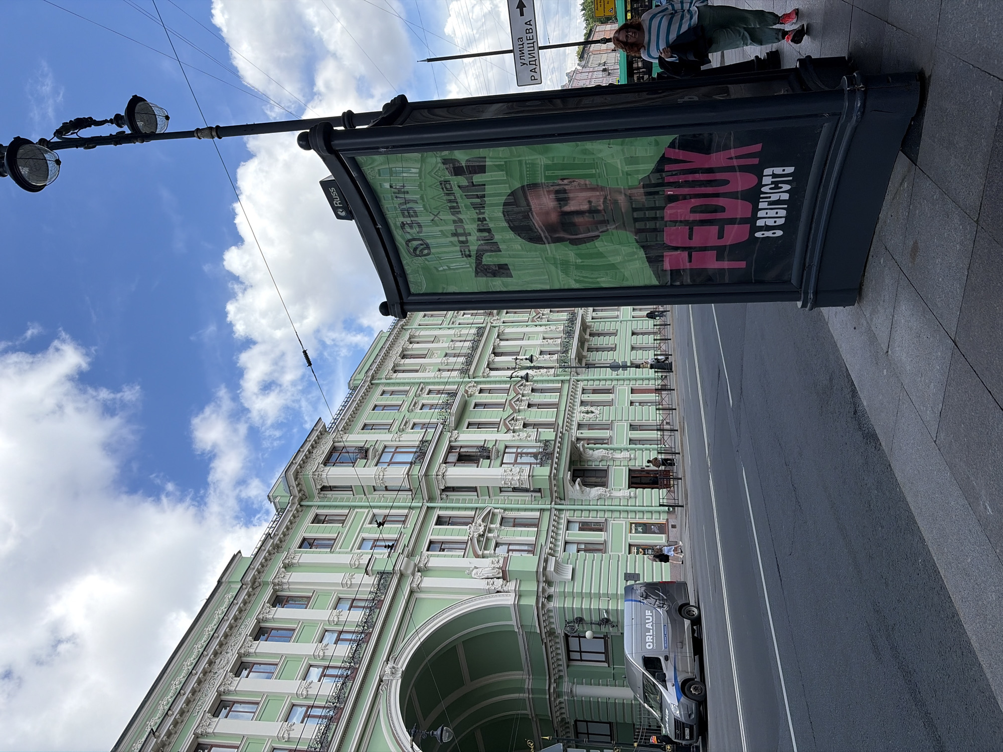 The image captures a street scene in Saint Petersburg, Russia, dominated by a light green ornate building with arched entrances. In the foreground, a large advertisement panel displays a portrait of a man and text in Russian, including "FEDUK" in large pink letters and "8 Августа" below it, indicating an event on August 8th. To the left, a grey van with "ORLAUF" branding is parked on the street. A street lamp with two globes stands to the right of the advertisement. Further down the street, a woman in a striped shirt and green pants walks away from the camera. A street sign with an arrow points to "УЛИЦА РАДИЩЕВА" (Radishcheva Street). The sky is blue with scattered white clouds.