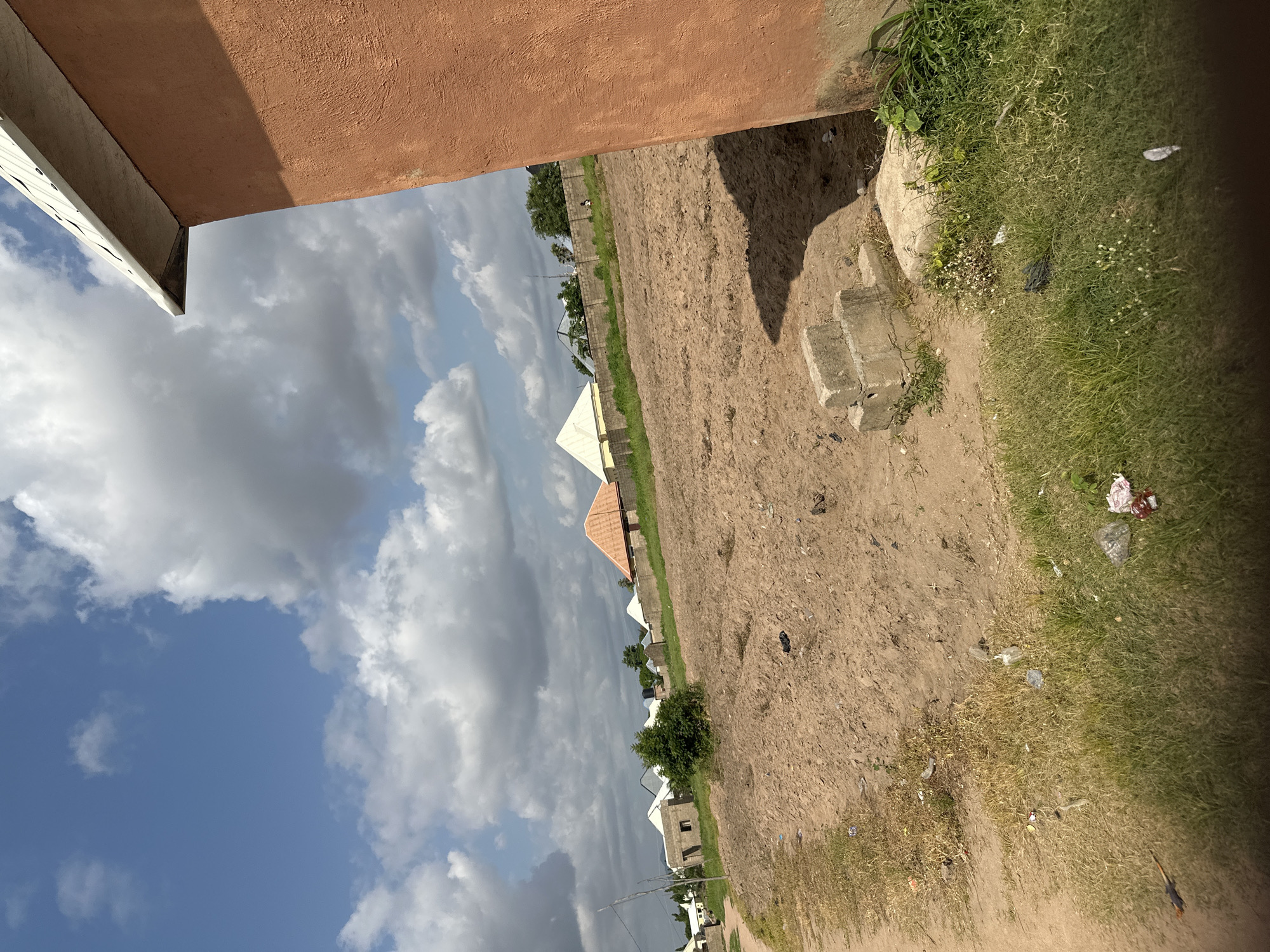 This media file, taken in Jalingo, Nigeria, depicts an outdoor scene under a cloudy sky. The perspective is from a low angle, looking up towards a reddish-brown building with a white soffit. To the left, a bright blue sky is filled with dramatic, large gray clouds, suggesting a dynamic weather system.
The main part of the image is a dirt and grass area. A dirt path runs diagonally through the frame, bordered by patches of green grass, some of which appear dry or sparse. Scattered on the ground are various pieces of litter, including plastic bags and wrappers, indicating a less than pristine environment.
In the middle ground, a line of buildings with pointed, light-colored roofs is visible, suggesting a residential or commercial development. Some of these buildings are under construction, with exposed brickwork visible on one. A low stone wall or fence separates this development from the dirt and grass area. Beyond this, more trees and buildings can be seen in the distance.
To the right of the frame, a concrete structure, possibly part of a step or foundation, is visible amongst the grass. The lighting suggests it is daytime, likely late morning or early afternoon, given the shadows. There are no discernible people in the frame, and no text is visible. The overall impression is one of an undeveloped or developing area on the outskirts of a town, with a mix of natural elements and human construction.