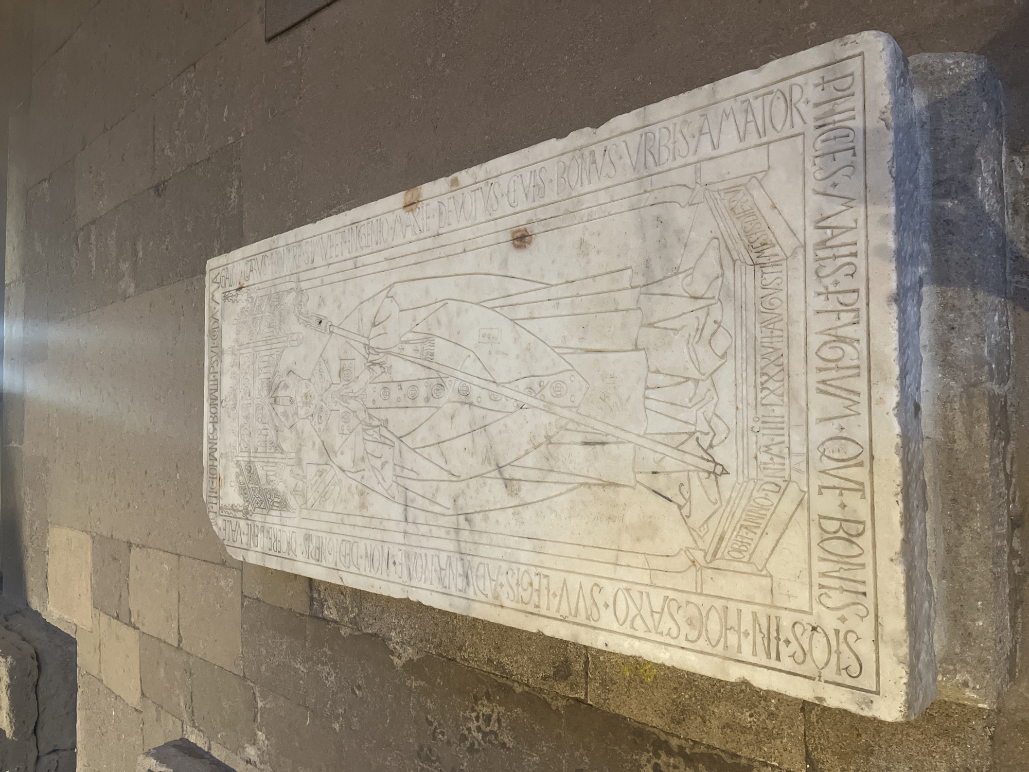 The image shows a long, rectangular white marble tombstone with carved inscriptions and a relief sculpture of a person in clerical or regal attire. The tombstone is set on a stone base and lies on a flagstone floor. The carving depicts a figure wearing a mitre and holding a crozier, suggesting it is a bishop's tomb. Surrounding the central figure are Latin inscriptions. The tombstone appears to be located in a historical building, possibly a cathedral or church, given the stone architecture visible in the background and the nature of the tomb itself. The lighting suggests an indoor environment with ambient light. The text is partially legible and seems to commemorate a historical figure with dates and titles. Based on its historical significance, this tomb is likely located in the former cathedral of Saint-Pierre in Villeneuve-lès-Maguelone, France, which was once the seat of the archbishops of Maguelone.