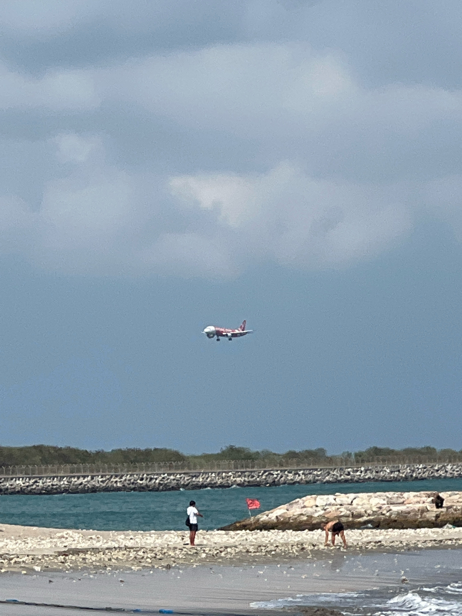 An airplane with red and white livery is flying in a gray sky above a beach. The plane is small in the frame, suggesting it is distant. Below the plane, the sea is a vibrant turquoise. A breakwater made of large stones and concrete forms a line across the middle ground of the image. On the beach in the foreground, people are visible. One person stands near the water's edge, wearing dark shorts and a white t-shirt, facing away from the camera. Another person is bent over on a rocky section of the beach, wearing dark shorts. A red flag is planted on the rocky area. The beach is covered with small white stones and debris. The overall impression is a beach scene under a cloudy sky.