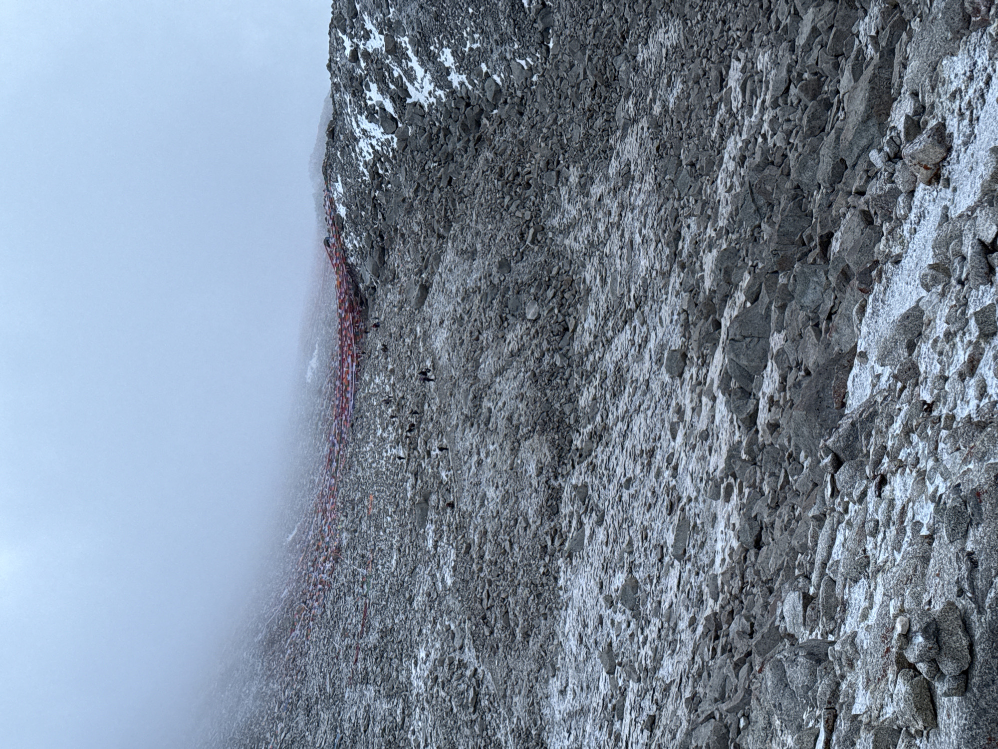This vertical image depicts a rugged and steep mountainside, partially enveloped in dense mist or clouds, suggesting a high-altitude environment. The terrain is dominated by loose grey scree, angular boulders, and scattered patches of snow, indicating cold conditions.
A prominent feature is a long string of colorful prayer flags, predominantly red, orange, and white, draped horizontally across the mid-left section of the image. These flags follow a path or ridge along the mountain. Below and alongside these flags, several small, dark human figures are visible. They appear to be a line of climbers or trekkers ascending the challenging slope.
The activity taking place is mountain climbing or high-altitude trekking. The climbers are moving in a single file, their small scale emphasizing the vastness and steepness of the mountain.
The weather is overcast and misty, limiting visibility in the upper parts of the scene and creating a diffused, soft light. No specific time of day is discernible, but it appears to be daylight. There is no visible text in the image.
While the visual evidence strongly suggests a high mountain range like the Himalayas, the user-provided location context for this media file is "Parga town, China."