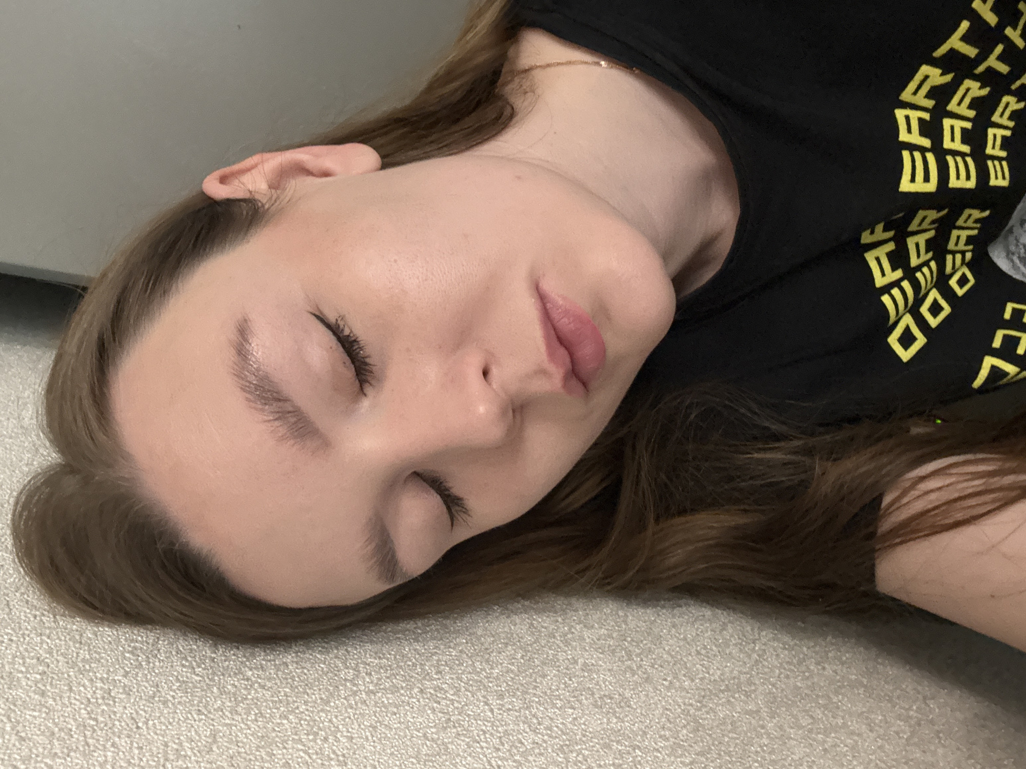 A close-up, slightly overhead shot captures a person, likely female, lying down indoors, appearing to be asleep or resting. The individual is positioned diagonally across the frame, with their head resting on a light-colored, textured surface, resembling a light grey or beige carpet. Their eyes are closed, and their facial expression is peaceful. Visible features include a subtle application of eyeliner and defined eyebrows. A thin, golden chain necklace adorns their neck.
The person has medium brown hair, which is fanned out around their head. They are wearing a black t-shirt. The shirt features yellow, stylized block letters, clearly showing the word "EARTH" repeated several times, and other partial letters.
The background consists of a plain, light-colored wall or surface. The lighting is soft and even, consistent with an indoor environment. The overall mood of the image is one of calm and repose. The location context provided for this media file is Izhevsk, Russia.