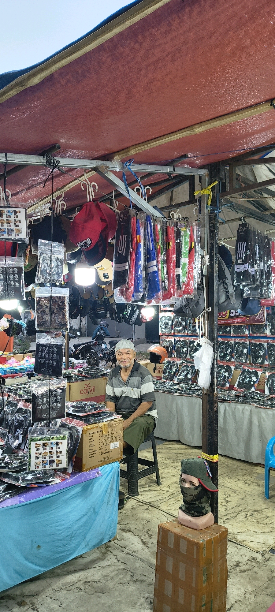 The image depicts an outdoor market stall in Trusmi Wetan, Indonesia. A middle-aged man with a light complexion, wearing a striped t-shirt and green pants, is seated on a black stool. He is looking directly at the camera and smiling. The stall is filled with merchandise, including hats, bags, and what appear to be motorcycle accessories. The stall is covered by a red tarp, and various items are displayed on racks and tables. In the background, motorcycles are visible, suggesting this is a busy marketplace. A mannequin head wearing a camouflage mask and a green cap is placed on a cardboard box to the right of the seated man. The ground appears to be concrete with a mottled pattern. The overall impression is a lively street market scene.