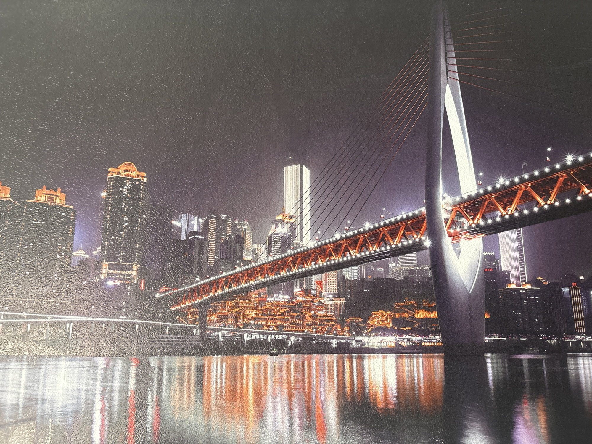 The image captures a vibrant city skyline at night, characterized by a large, illuminated cable-stayed bridge spanning a body of water. The time of day is night, with countless artificial lights from buildings and the bridge creating a luminous scene.
The central feature is a modern bridge with a prominent, elegant white/grey pylon that splits into two arches near its top. Numerous cables extend from this pylon, supporting the bridge deck, which is a truss-like structure adorned with bright white lights along its length and warm orange lighting beneath. The lights create striking starburst effects.
On both banks of the river, a dense urban landscape unfolds. Numerous high-rise buildings and skyscrapers, with many illuminated windows, form a backdrop. A particularly notable feature on the left bank, beneath the bridge, is a multi-tiered complex of traditional-style architecture, brightly lit with warm orange and yellow tones, reminiscent of the Hongyadong complex. The entire cityscape, including the bridge and buildings, is vividly reflected on the calm surface of the water, suggesting clear and still weather conditions.
No people are visible in the image, and there is no discernible text. The scene conveys a sense of urban grandeur and nocturnal activity. Based on the distinctive bridge and architectural features, the location is identifiable as Chongqing, China, specifically featuring the Qianximen Bridge and the Hongyadong complex. The image itself has a noticeable textured, slightly coarse quality, suggesting it might be a print on canvas or a similar material.
