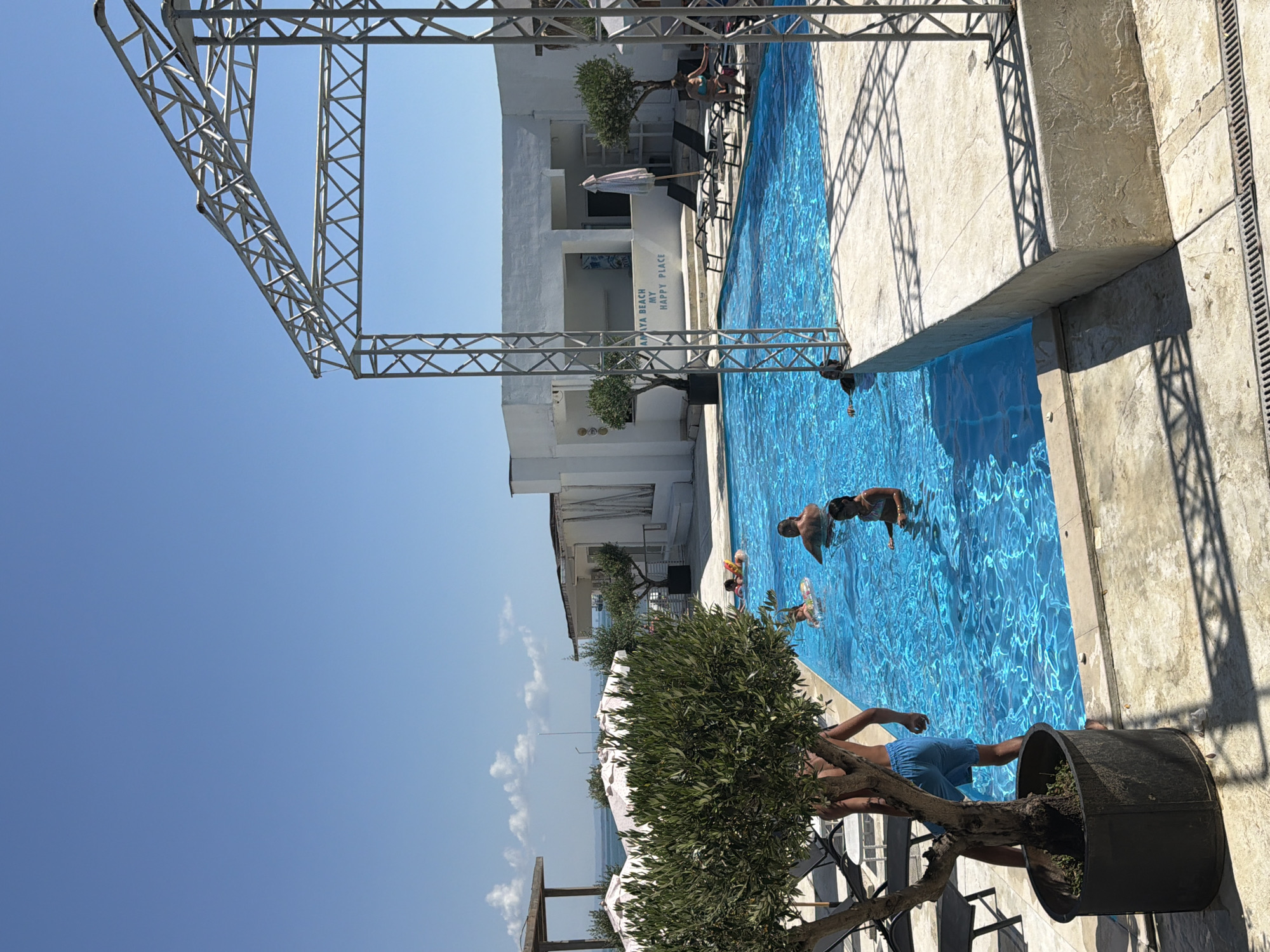 The image depicts a sunny outdoor scene at a swimming pool, likely part of a resort or beachfront establishment. In the foreground, to the left, a potted olive tree with dense green foliage stands prominently. To the right, a concrete structure, possibly a stage or platform, extends towards the pool. The pool itself is filled with vibrant blue water and several people are swimming and relaxing. A man and a young girl are visible in the water on the right side of the pool. Further back, other individuals, including children, are enjoying the water. Beyond the pool, a white building with a covered outdoor area and umbrellas suggests a hospitality venue. The background shows the sea and a clear blue sky. The text "AMAYA BEACH MY HAPPY PLACE" is visible on the white building, indicating the name of the location. The overall impression is one of leisure and vacationing. This scene is consistent with a location in Burgas, Bulgaria, known for its coastal resorts and beaches.