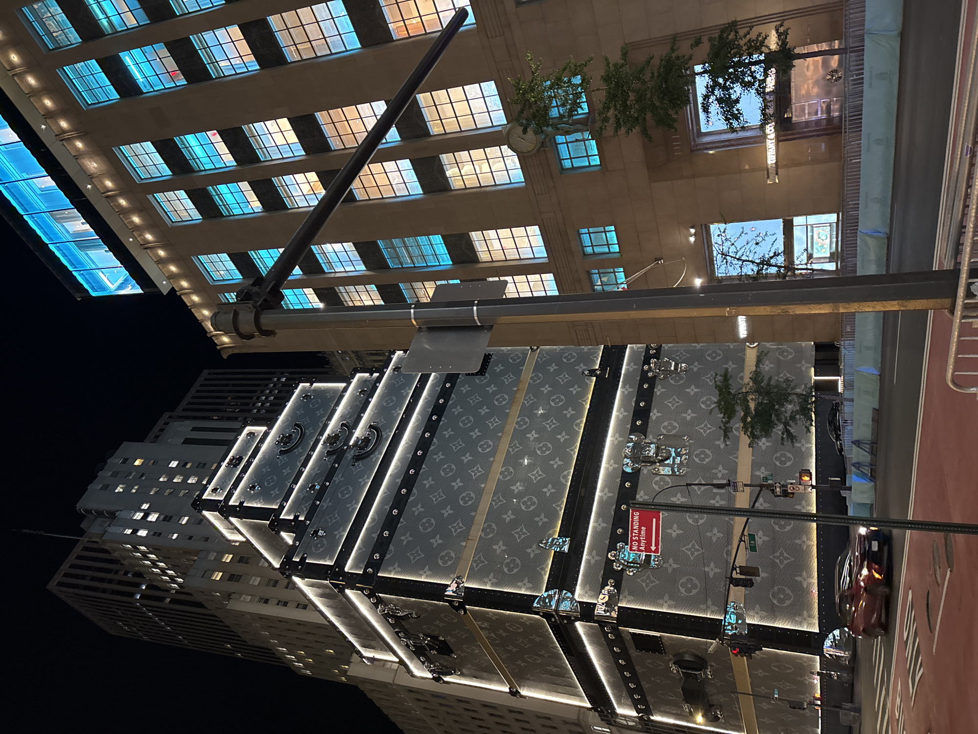 This nighttime urban scene captures a towering building adorned with a unique facade designed to resemble stacked Louis Vuitton trunks. The trunks, prominently featuring the brand's iconic monogram pattern, are dramatically lit with bright white LED strips outlining their edges and hardware, creating a striking visual spectacle.
In the background, other tall city buildings are visible. One prominent building to the right features numerous windows, some glowing with warm yellow light and others with cool blue light. Another building partially visible to the far left also displays many illuminated windows. Overhead, some green trees are seen growing on building ledges or in planters.
At the bottom of the image, a street is visible, with a red car partially entering the frame. The street has a distinct red-painted lane, possibly for bicycles or buses. Street furniture, including a large pole with traffic lights and various street signs, punctuates the foreground.
The time of day is clearly night, as indicated by the dark sky and the reliance on artificial lighting from the buildings and street. The overall impression is one of a bustling, high-end commercial district.
Visible text includes the prominent Louis Vuitton monogram on the decorative trunks. A red rectangular sign clearly states "NO STANDING ANYTIME." A green street sign, although partially obscured, shows "57 ST" and a smaller "W" (for West 57th Street). Another building in the background, visible in one of the provided crops, has "TIFFANY & CO" written vertically. These cues strongly suggest the location is New York City, specifically around the intersection of 57th Street and Fifth Avenue. There are no people clearly visible in the image.
