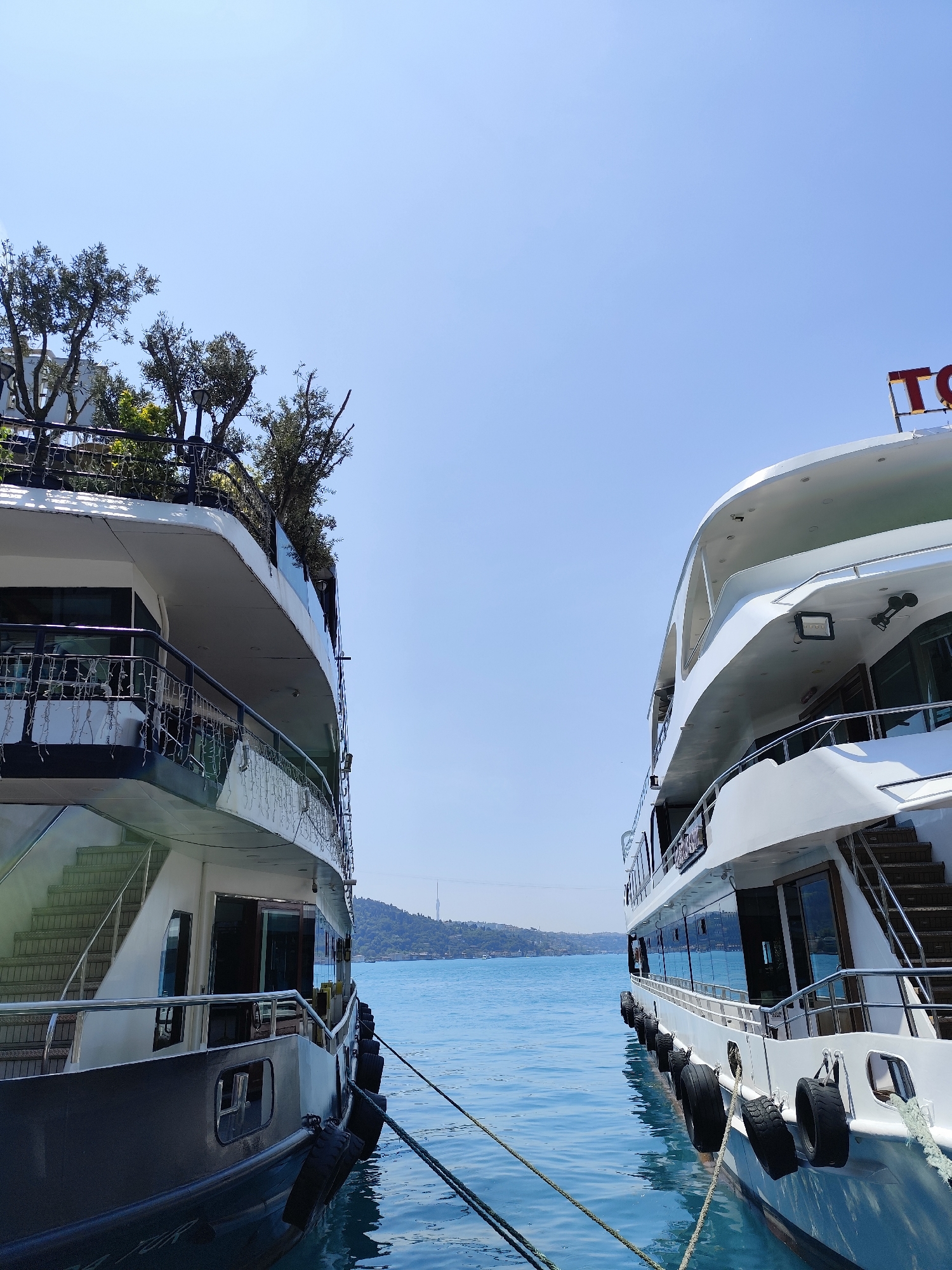 The image captures a sunny daytime scene in Beşiktaş, Turkey, showcasing two large, multi-decked boats docked side-by-side, framing a view of the Bosphorus Strait. The clear blue sky indicates excellent weather.
The boat on the left has a white upper structure over a darker hull. It features multiple levels with balconies and railings, from which decorative string lights hang. Its most prominent detail is the lush green foliage and trees growing on its top deck, creating a distinctive garden-like appearance.
The boat on the right is predominantly white, mirroring the multi-level design with numerous windows and railings. A partial red sign, displaying the letters "TO...", is visible on its upper deck. Both vessels are secured to the dock with thick ropes, and black rubber fenders line their sides for protection.
Between the two boats, a calm expanse of blue-green water stretches towards the horizon. In the far distance, a green, hilly landscape is visible, topped by what appears to be a suspension bridge (likely one of the Bosphorus bridges) and a tall, slender structure, possibly a communication tower. The scene conveys a sense of peaceful maritime activity under a bright, cloudless sky. No people are distinctly visible.