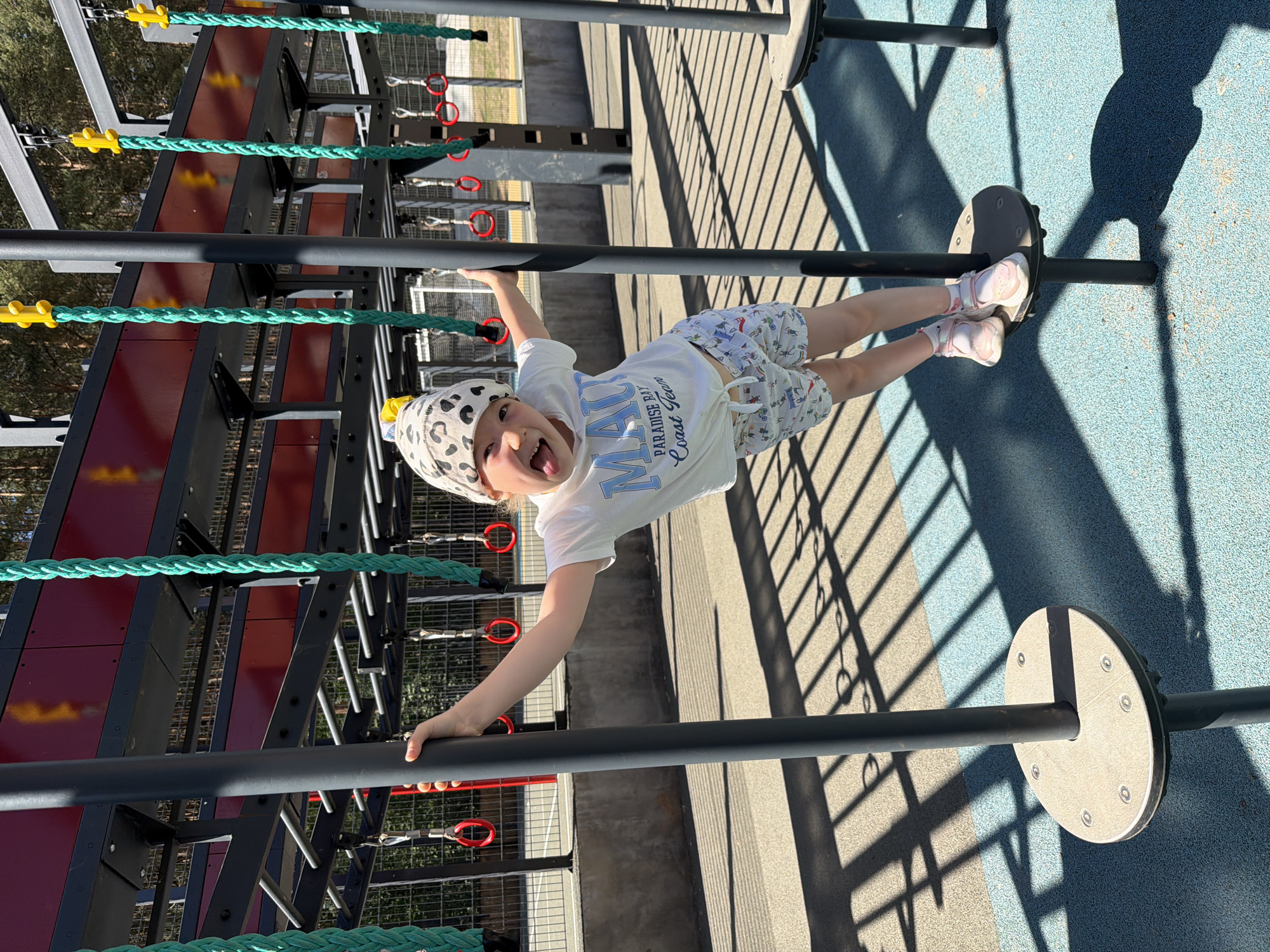 A child wearing a white t-shirt, patterned shorts, and a patterned hat is hanging upside down on a playground structure. The playground equipment consists of horizontal bars, ropes, and platforms. The ground is a blue and tan material. Shadows from the playground structure are cast on the ground. The location is not identifiable as Izhevsk, Russia from the given image.