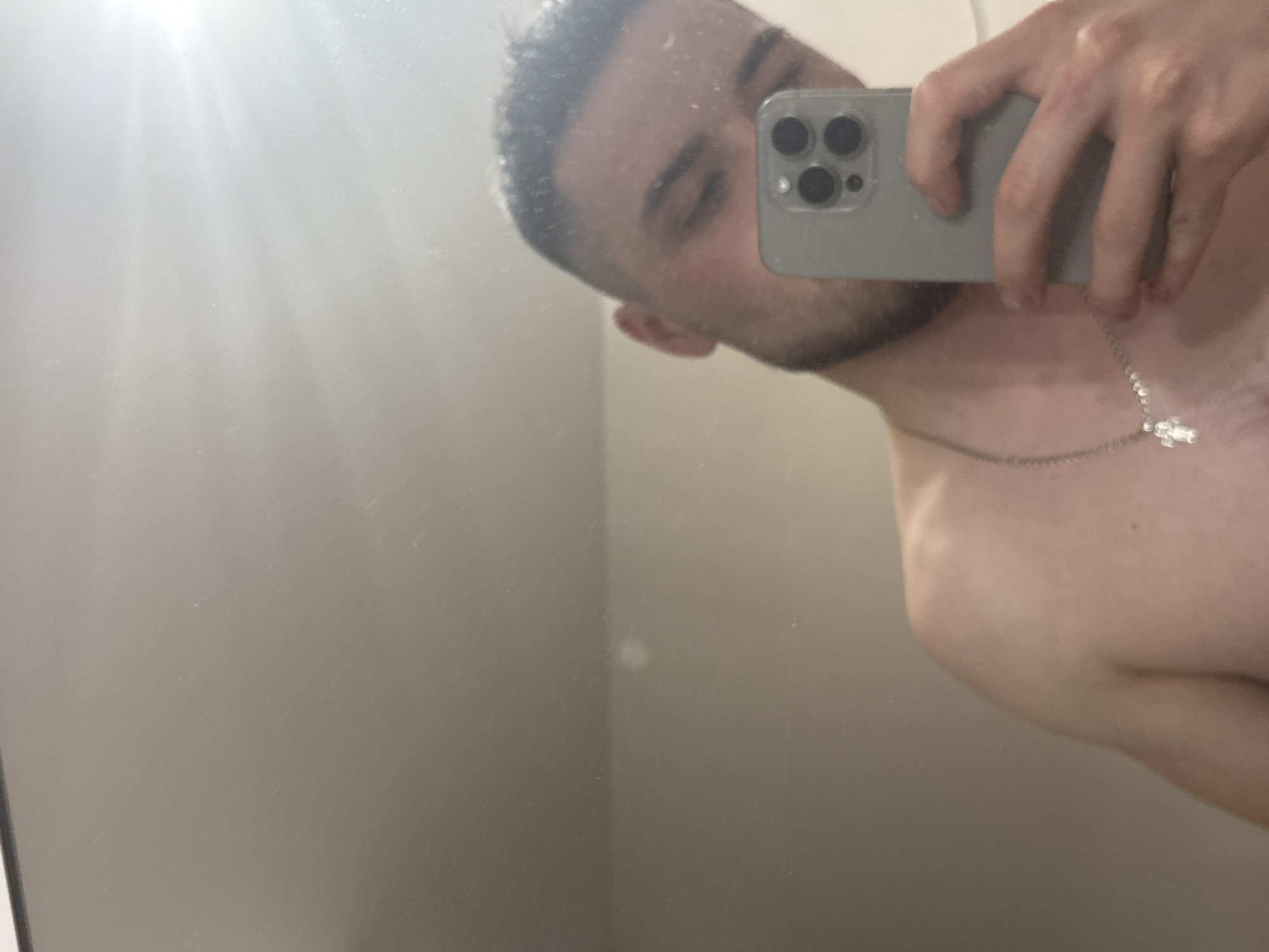 The image is a mirror selfie featuring a young man captured from the chest up. He holds a light grey smartphone, likely an iPhone with its distinctive three-lens camera array, in his right hand to take the picture. The man has short dark hair and a beard, and his head is slightly tilted to his right. He is shirtless and wears a silver chain necklace with a small, clear or white pendant resting on his chest. His eyes are partially closed or cast downwards, giving him a neutral expression.
The setting is indoors, with a light-colored wall reflected in the mirror as the background. A bright light source, possibly a window or an overhead fixture, creates a prominent lens flare in the upper left corner of the image. The mirror surface appears to have some dust or smudges, contributing to the casual aesthetic of the selfie. No specific location cues, such as text or distinctive architectural features, are visible within the image to confirm the provided context of Perm, Russia. The time of day is not discernible from the indoor setting.