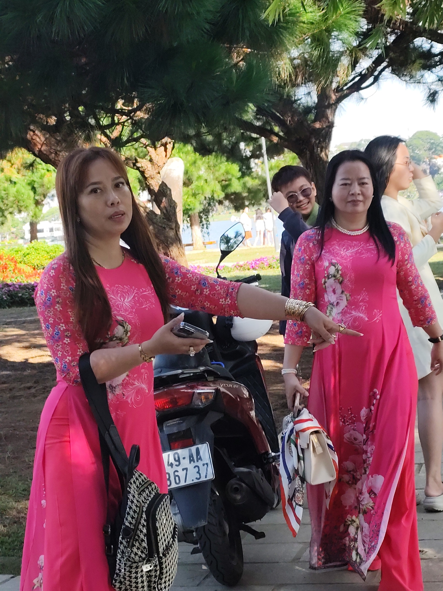 In Da Lat, Vietnam, a group of women are posing outdoors. The woman in the foreground, on the left, wears a bright pink dress with a floral pattern and a black and white patterned backpack. She is looking to the side and holding a cell phone in her right hand. To her right, another woman in a similar pink floral dress stands with a small beige handbag and a patterned scarf. Behind them, a younger person wearing sunglasses and a dark jacket is smiling. Further in the background, another woman in a light-colored outfit is visible. They are standing on a paved area with trees and lush greenery in the background, suggesting a park or garden setting. A motorcycle is parked to the right of the woman in the foreground.