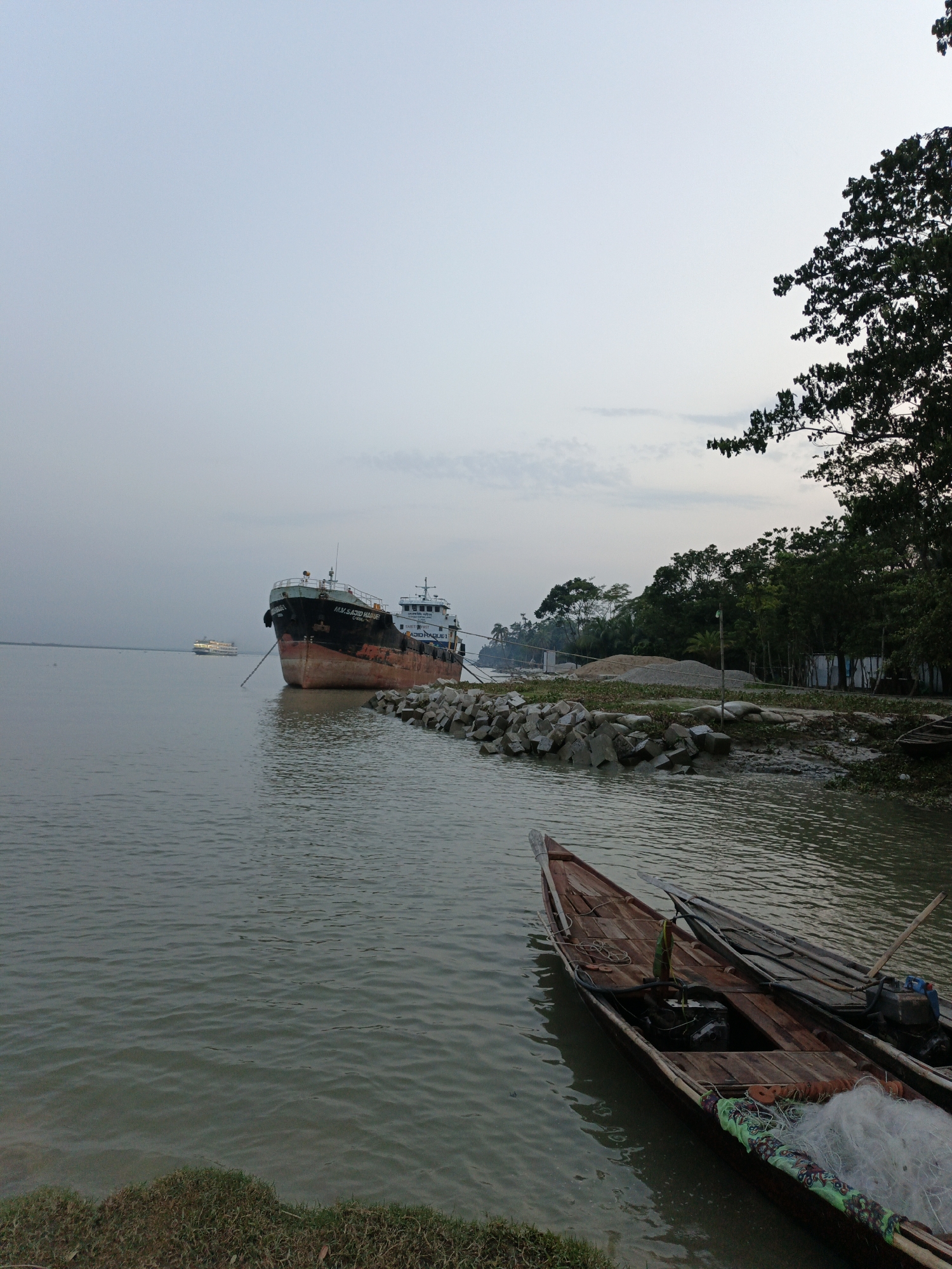 The image depicts a waterfront scene in an unknown city. A large cargo ship is moored offshore, with a smaller passenger ferry visible in the distance. Along the shore, there is a breakwater constructed of large concrete blocks. A pile of aggregate or construction material sits near the waterfront, adjacent to some small buildings. In the foreground, two wooden boats are moored to the shore, containing fishing nets and small outboard motors. A tree line is visible on the right side of the image. The sky is overcast.