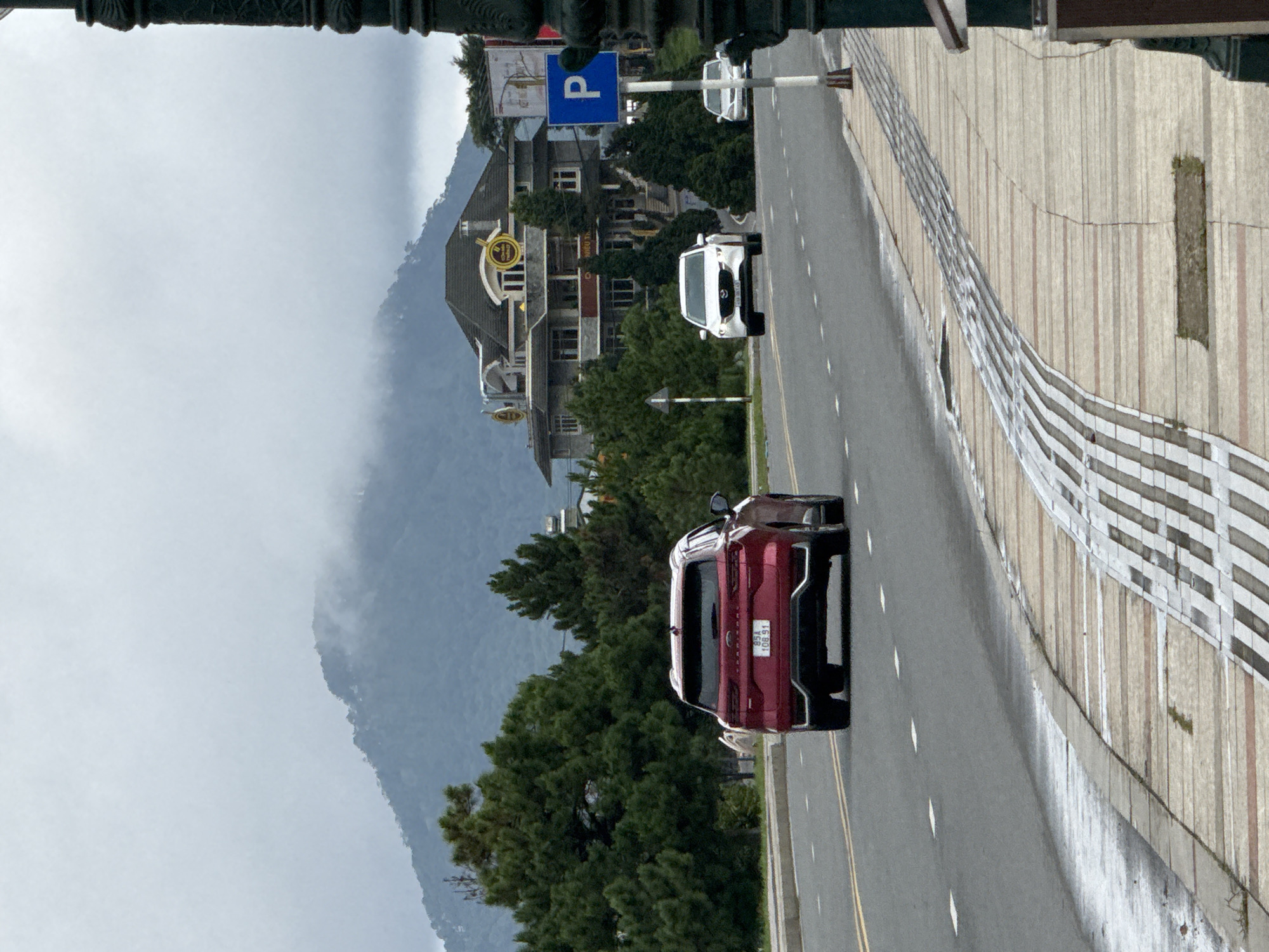 The image depicts a street scene in Xuân Hương Lake, Đà Lạt, Vietnam, on an overcast day. A red SUV is in the foreground, driving on a paved road with visible white lane markings. To the right of the road, a textured sidewalk with a brick pattern and decorative metal grate is visible. Beyond the road, several cars are driving, including a white sedan in the middle ground and another white car further in the distance. On the right side of the image, a dark decorative lamppost is present. In the background, a large, multi-story building with a distinctive roofline and signage is situated amongst lush green trees. Further in the distance, a misty mountain range is shrouded in clouds, indicating the mountainous terrain of the region. A blue parking sign with a white "P" is posted on a pole near the building. The overall impression is a serene, somewhat subdued urban environment integrated with natural landscapes.