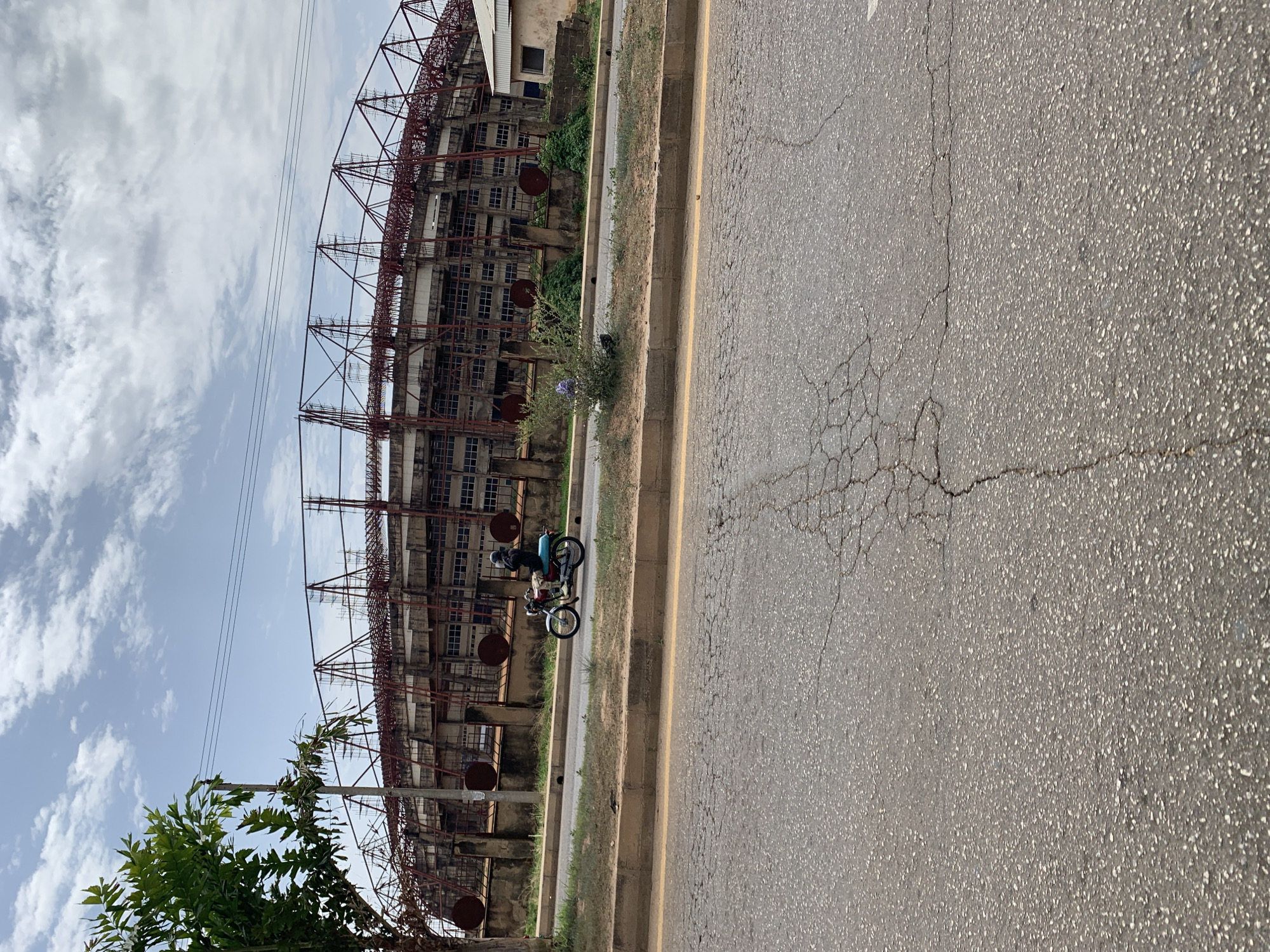 The image depicts a weathered asphalt road in front of a large, unfinished concrete and steel stadium structure, identifiable as the New Stadium in Nigeria due to its design. The road shows signs of cracking and wear. A motorcyclist is seen riding adjacent to the stadium on the right side of the road. The sky is visible in the background with sparse clouds. Vegetation grows sparsely along the edge of the road.