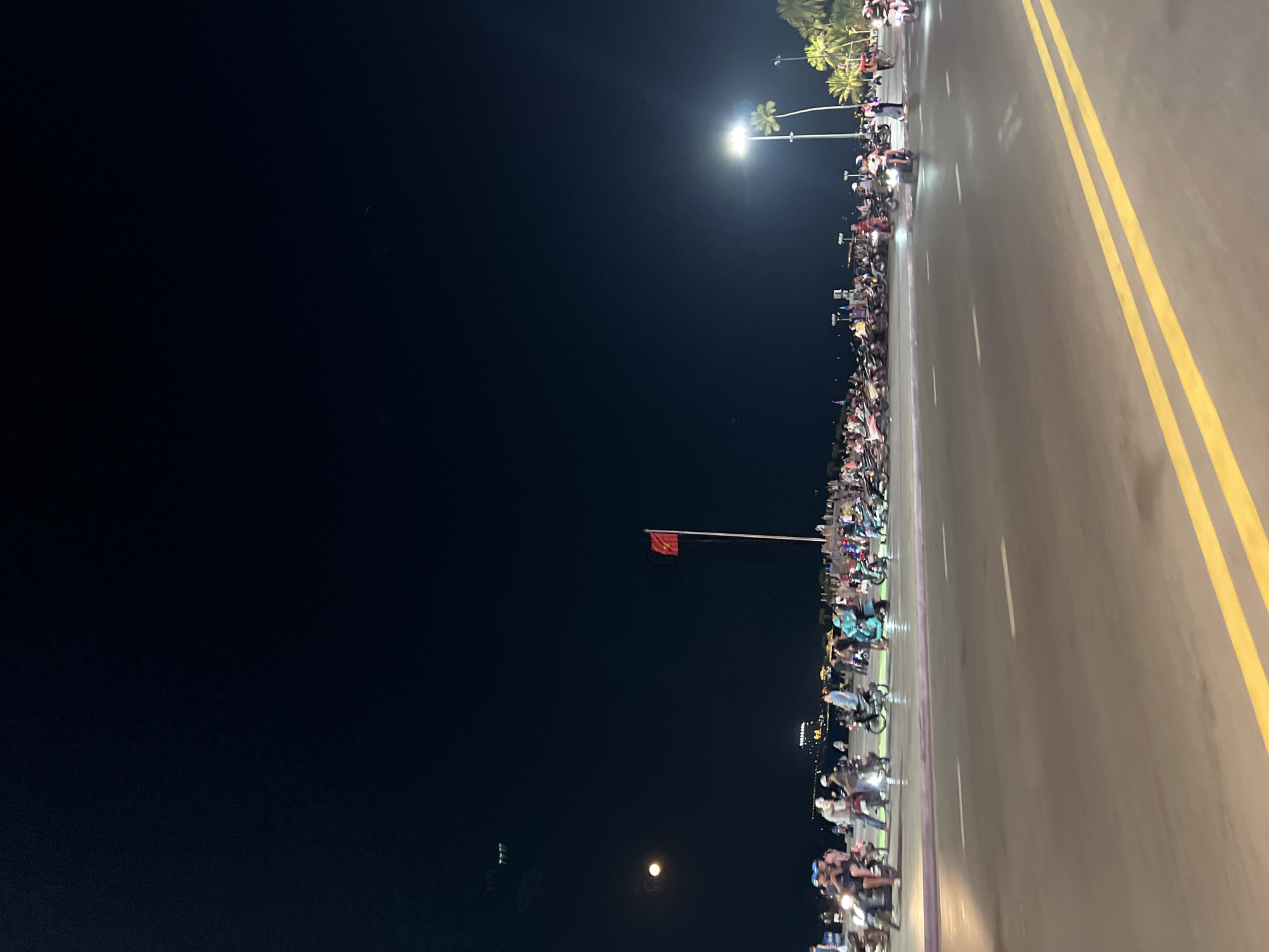 The image shows a street scene at night in Nha Trang, Vietnam. A large crowd of people on motorbikes fills the right side of the frame, lining the street. Double yellow lines mark the road. Streetlights illuminate the area. The Vietnam flag is visible on a tall pole. In the distance, palm trees are lit up. The night sky occupies the left side of the frame.