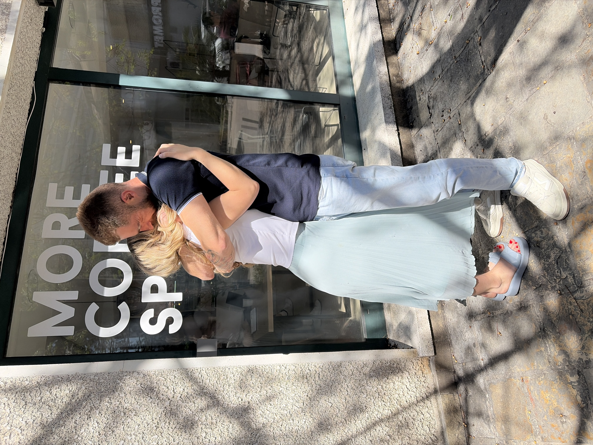 A man and a woman are embracing on the sidewalk in front of a building with a large glass window. The window has the words "MORE COFFEE SPACE" printed on it. The man is wearing a navy blue t-shirt and light blue jeans, with white sneakers. The woman is wearing a white tank top and a light blue pleated skirt, with blue platform sandals. The sidewalk is made of stone, and there are shadows cast on it. The location is Budva, Montenegro.