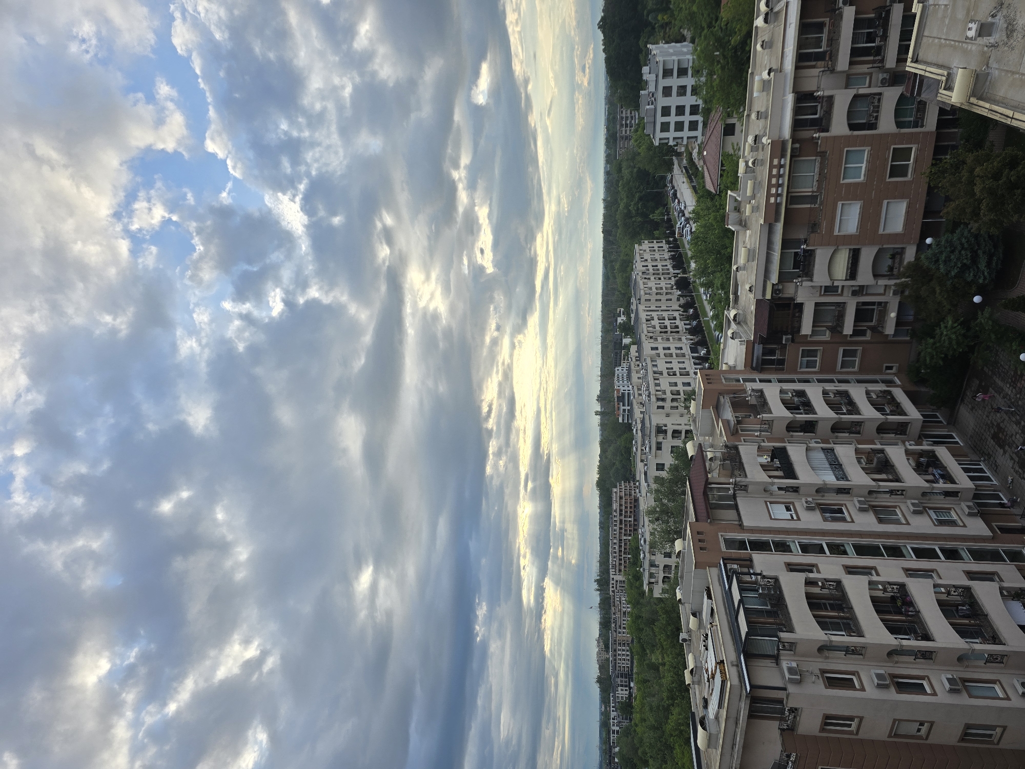 The image shows a cityscape with apartment buildings on the right side, and a cloudy sky on the left. There are tall buildings with multiple floors and balconies. A line of trees is visible along the side of the buildings. The sky is filled with clouds of varying shades of gray and white, with some blue sky peeking through. The location appears to be a city.