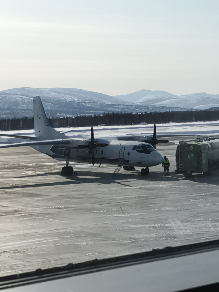 At an airport in Сокол, Russia, a white twin-engine turboprop aircraft, likely an Antonov An-24 type, is parked on a partially snow-covered tarmac. A single ground crew member, wearing a high-visibility yellow-green jacket and dark trousers, stands near the aircraft's nose. A green fuel tanker truck is positioned adjacent to the aircraft's front section. An open boarding ladder is deployed at the forward-left passenger door of the aircraft. The tarmac shows visible tire tracks and scattered patches of snow and ice. In the background, a dark treeline fronts a range of snow-covered mountains or large hills, beneath a clear, bright sky. The image is framed from an elevated indoor perspective, with a white window frame visible along the bottom edge.