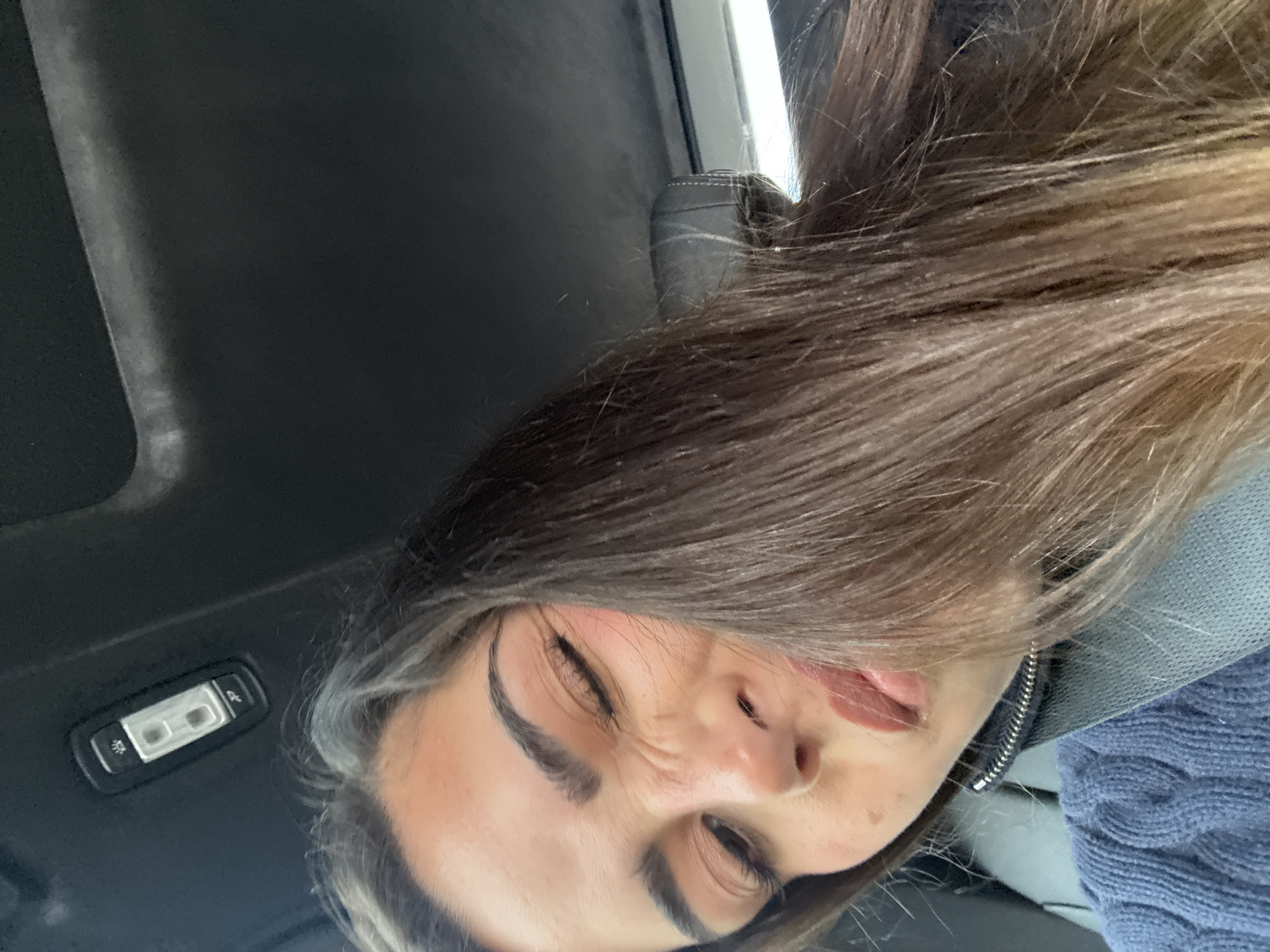 A woman with brown hair and fair skin is shown in a close-up selfie taken from a low angle. She is wearing a blue sweater and a seatbelt, and her hair is partially obscuring her face. Her eyes are squinted and she has her tongue slightly sticking out. The interior of a car is visible, including the dark gray ceiling and a car light fixture. There is no information available to determine if the picture was taken in the city of Valle de Chalco Solidaridad, Mexico.