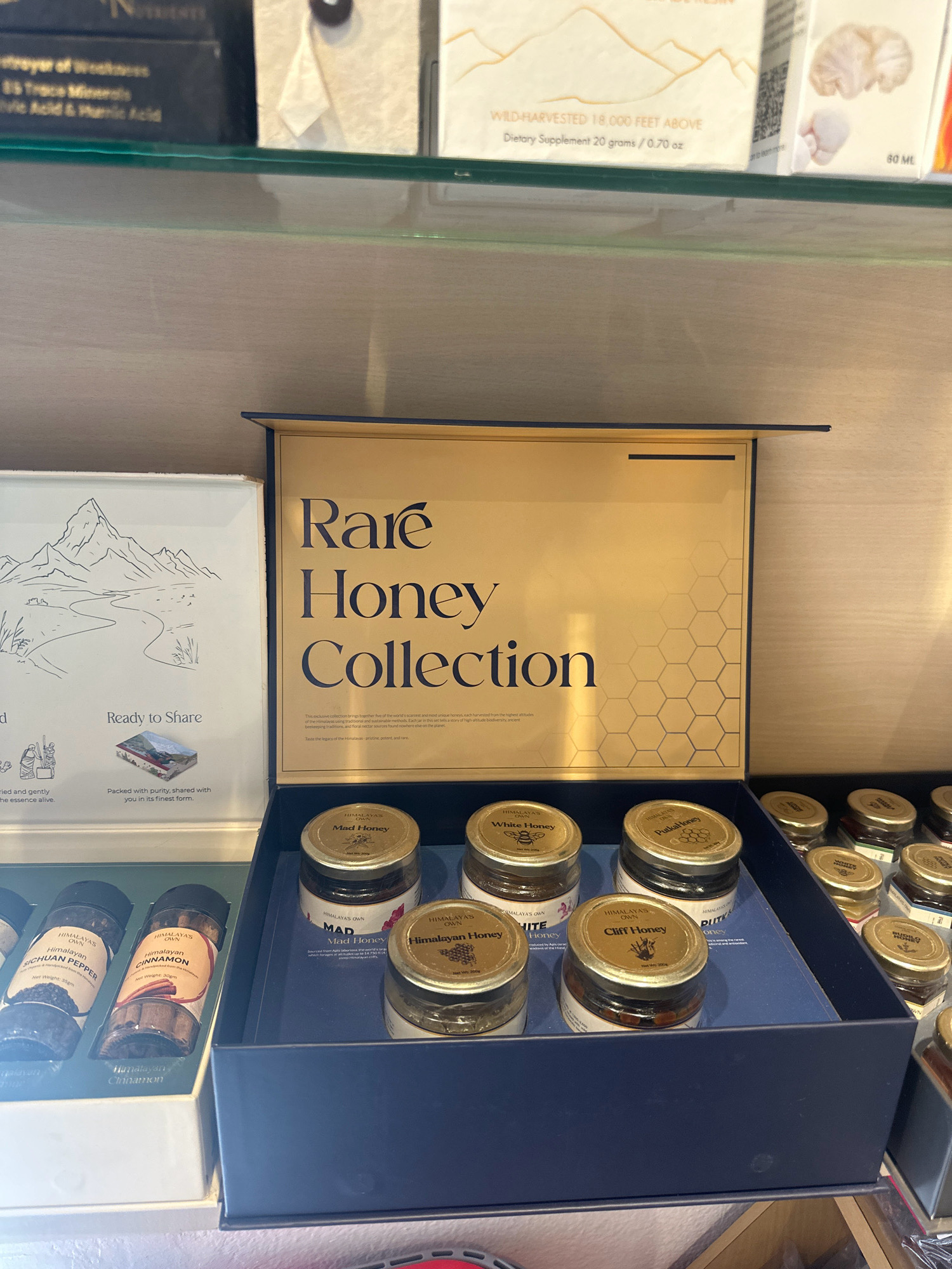 An overhead shot shows a blue box containing a collection of honey jars on a shelf. The box is open, revealing a gold-colored interior with a honeycomb pattern. Inside the box, six small jars of honey are arranged in two rows. The labels on the jars indicate various types of honey, including "Mad Honey," "White Honey," "Pukka Honey," and "Cliff Honey." In the foreground, to the left of the honey box, are two cylindrical spice jars, one labeled "HIMALAYAS OWN SICHUAN PEPPER" and the other "HIMALAYAS OWN CINNAMON." The background shows shelves with other products, including boxes and what appear to be larger jars or bottles. The lighting suggests an indoor setting, possibly a store or display. The image does not provide any specific location details within the city of Lalitpur, Nepal.