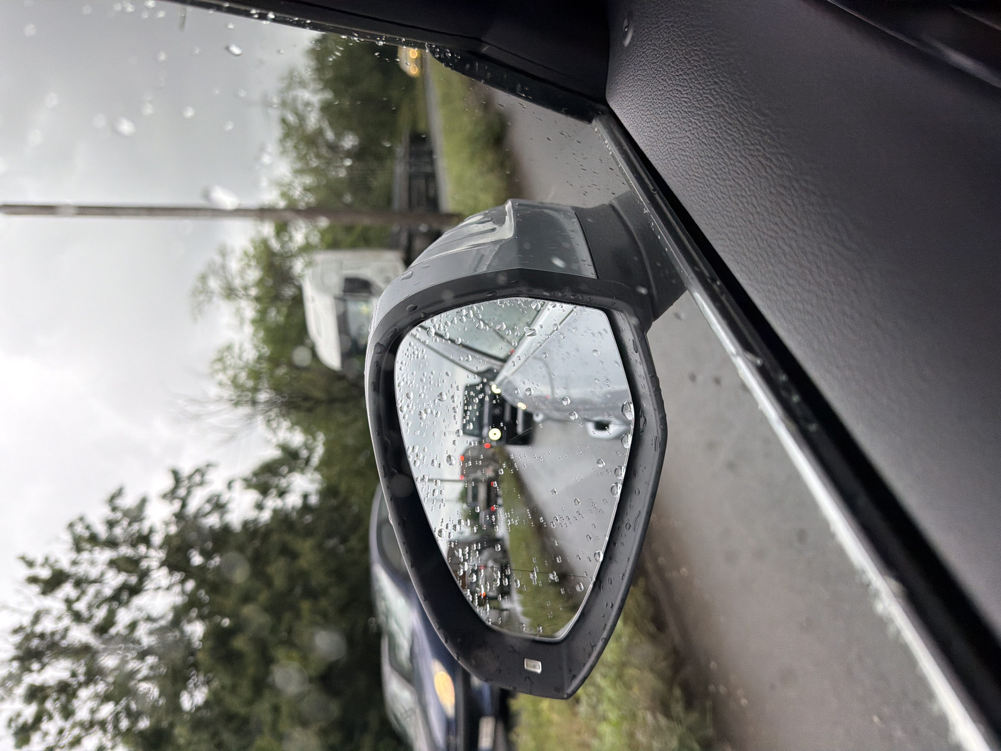 The image depicts a car's side mirror reflecting a traffic jam on a road. The mirror is covered in raindrops, obscuring the view to some extent. In the reflection, several cars and a white van are visible, with their red brake lights illuminated, indicating stopped or slow-moving traffic. Outside of the mirror, the background shows trees and a roadside environment. The perspective is from inside the car, looking out through the side window and mirror. The location in Burgas, Bulgaria cannot be confirmed from the image alone.