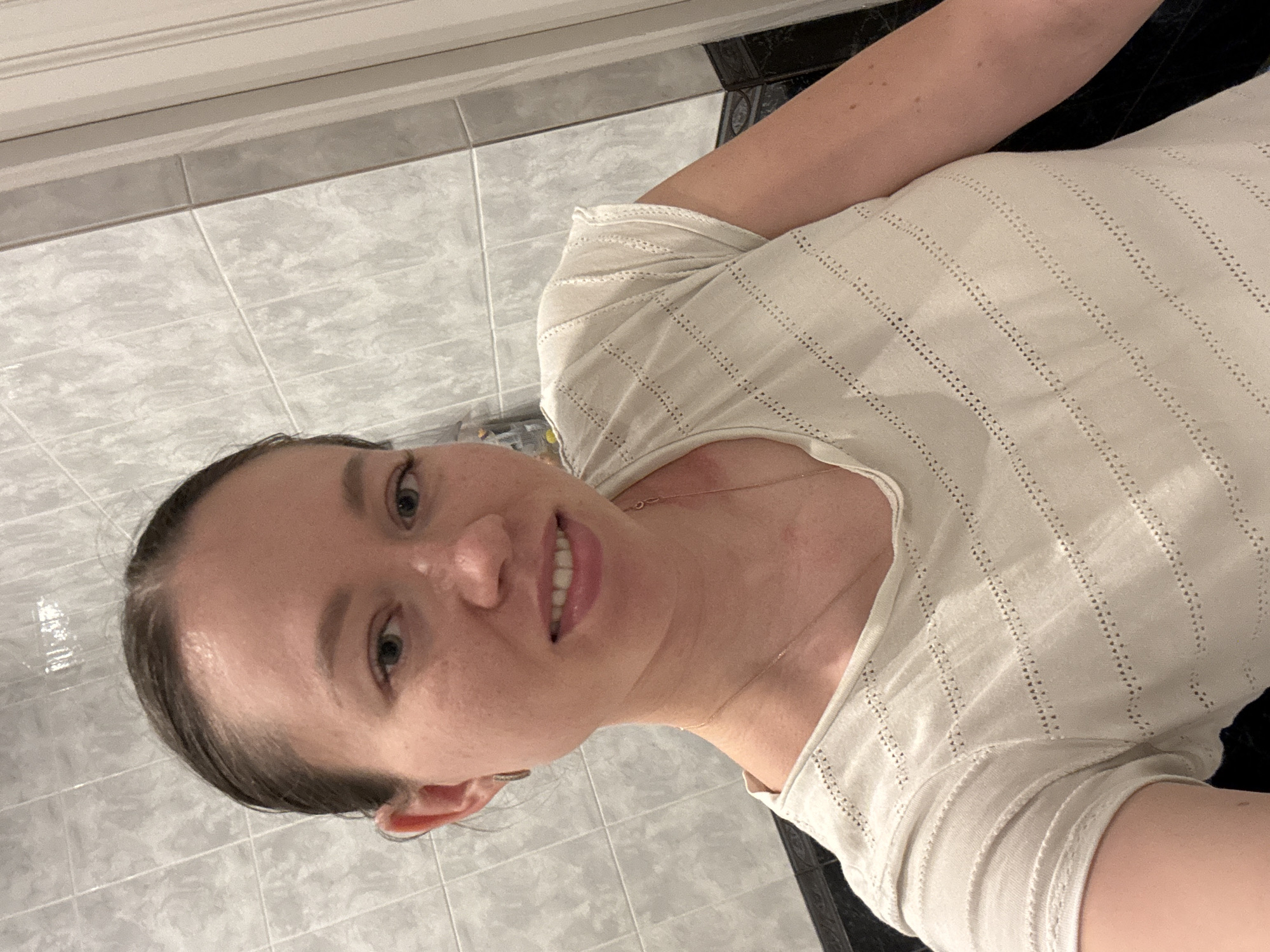 The image depicts a female subject in an indoor environment, identified as being in the city of Ферма, Russia. The woman, appearing to be in her 20s or 30s, is positioned centrally, facing the camera directly with a slight smile, revealing her upper teeth. Her dark hair is pulled back from her face. She has fair skin with visible freckles on her cheeks and nose. She is wearing a light beige or off-white short-sleeved top featuring horizontal perforated patterns. A thin, gold-colored chain necklace is visible, as is a small, gold-colored hoop earring in her left ear. The background consists of a wall covered in light grey rectangular tiles with subtle marbling or texture. Below these, a darker, possibly black, decorative tile border with an intricate lighter pattern runs horizontally. To the right of the subject, a vertical light-colored door frame or trim is visible.