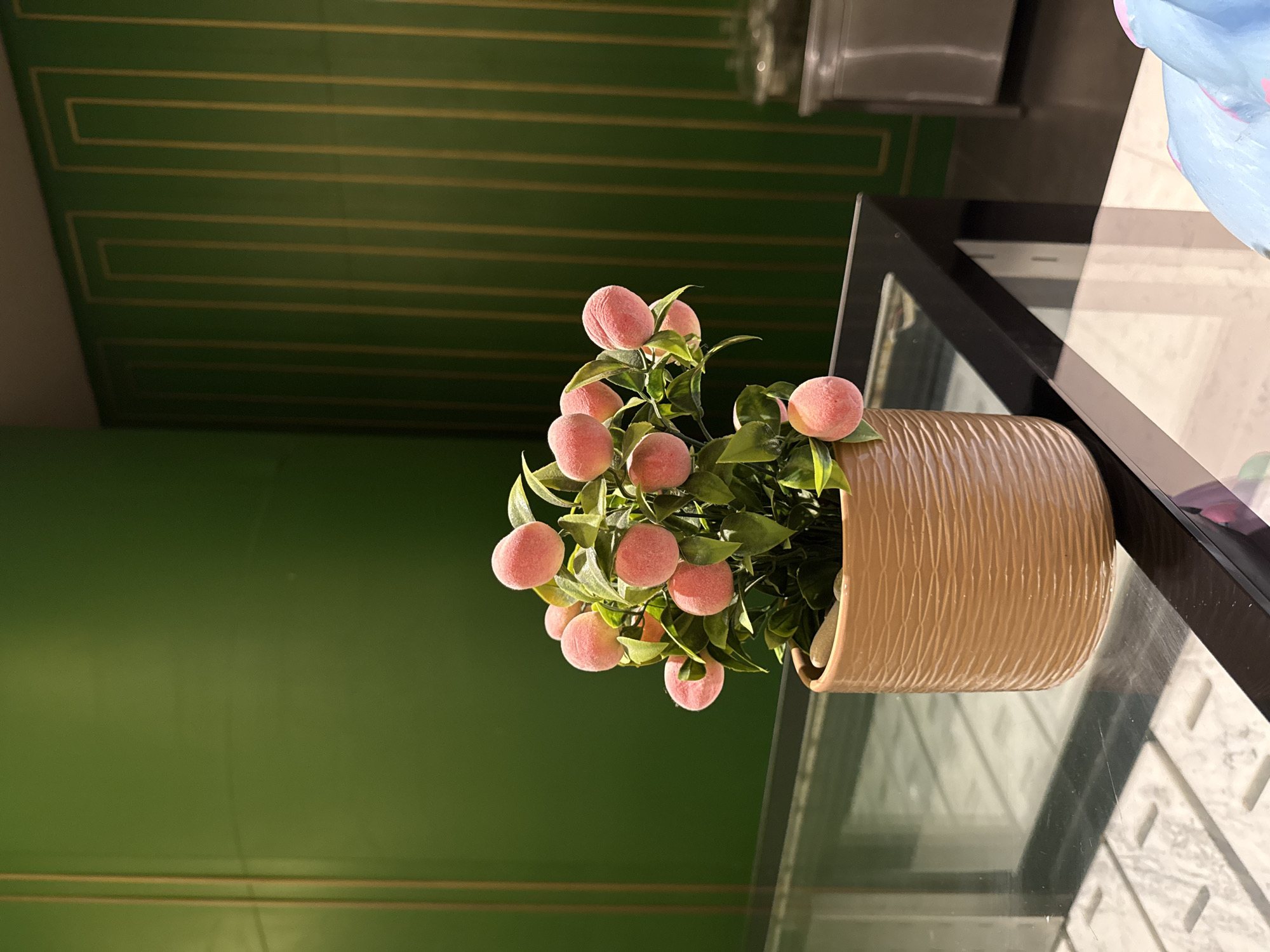 An artificial peach tree with several fuzzy pink peaches is displayed in a textured tan pot on a glass table. The table is set against a dark frame, and its surface reflects a grid-like pattern from below. The background features a vibrant green wall with decorative gold vertical stripes. In the far right background, a stainless steel appliance, possibly a refrigerator or display case, is visible. The scene appears to be indoors, likely in a commercial setting, though no specific location in Bodrum, Turkey is identifiable from the visual information.
