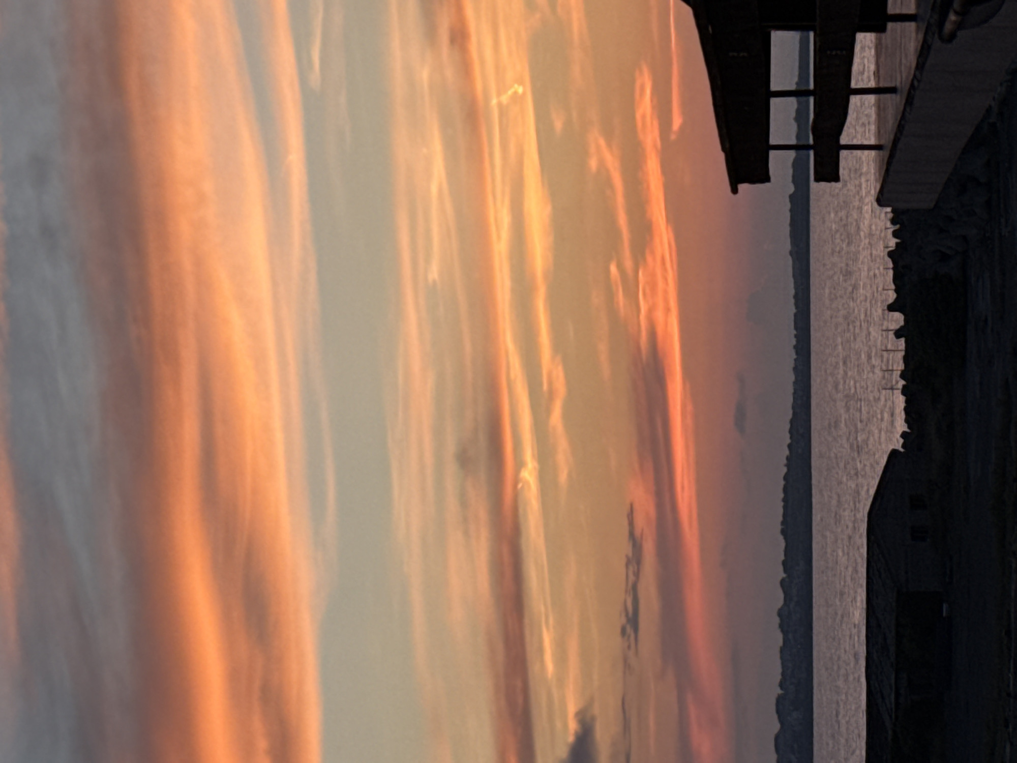 An image depicts a sunset over the Black Sea, with the sky displaying vibrant hues of orange, pink, and yellow. The water reflects the colors of the sky, creating a serene and picturesque scene. In the foreground, a dark, silhouetted structure with a cantilevered section is visible on the right, partially obscuring a low-lying building or shed on the left. The coastline of Sozopol, Bulgaria, appears as a dark, undulating line in the distance, suggesting a town or city. The image captures the tranquil beauty of a coastal sunset.