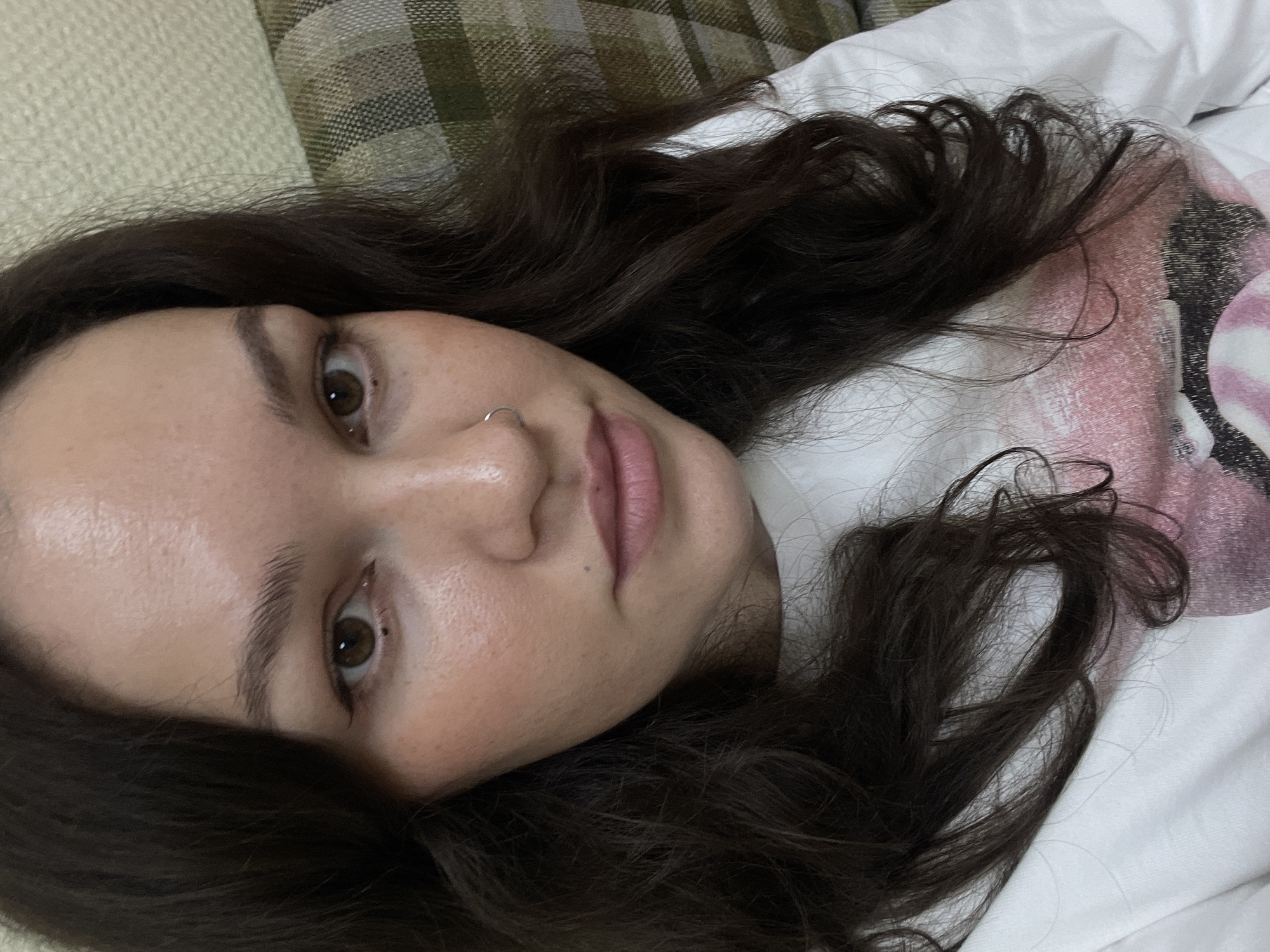A close-up selfie captures a young person lying down, looking directly into the camera. The subject has dark brown, wavy hair that spreads across a white pillow or bedding, and their head rests against a background of green and brown plaid fabric, possibly a blanket or upholstery.
The person's face is partially visible, showing one brown eye with a direct gaze, the nose, and the lips. A small, silver hoop nose ring is visible on their right nostril. Their skin appears fair with natural texture. They are wearing a white t-shirt, which has a faded pink and white graphic design on it, too indistinct to identify specific text or imagery.
The overall expression is neutral and relaxed. The activity taking place is a self-portrait, suggested by the close-up, angled shot. There are no explicit visual cues within the image to indicate the time of day or specific location, though the context provided specifies Izhevsk, Russia. No discernible text is visible in the image.