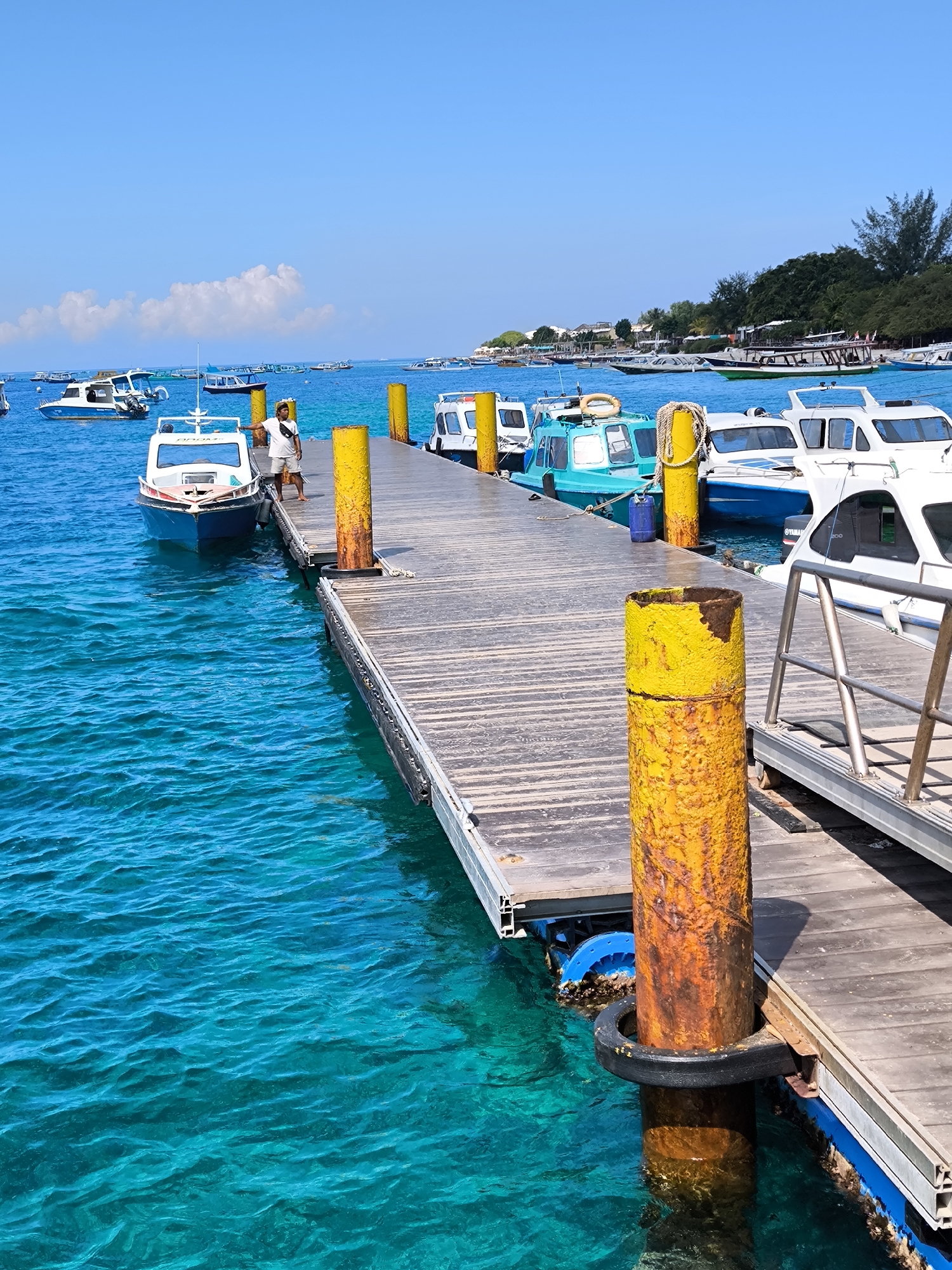 The photograph captures a dock in an unidentified coastal location in Indonesia, possibly in the city of Unknown. The foreground features a wooden dock with yellow bollards exhibiting signs of rust. Several boats are moored along the dock, including white and teal boats. A man wearing a white shirt and shorts stands on the dock next to a smaller blue boat. The clear, turquoise water surrounds the dock. In the background, a coastline covered in trees is visible with more boats moored in the water. The sky is clear blue with a few scattered white clouds.