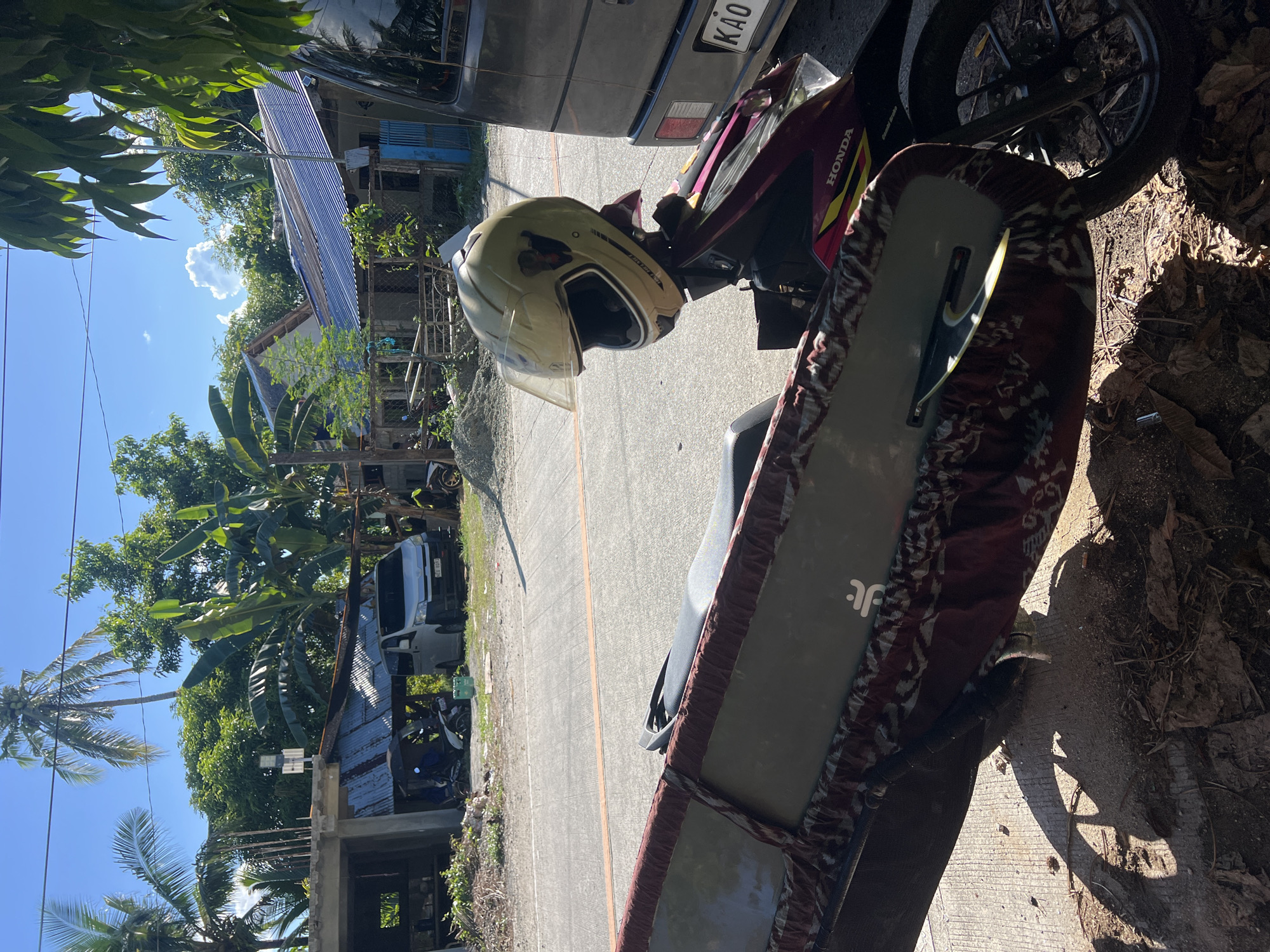 In General Luna, Philippines, a motorcycle is parked on the side of a paved road under a clear blue sky. A light yellow helmet rests on the motorcycle's handlebars. A long, flat object, likely a surfboard, is attached to the side of the motorcycle, covered in a patterned fabric. In the background, several residential structures with corrugated metal roofs and partially constructed buildings are visible, surrounded by lush green trees and palm trees. A white van is parked in front of one of the houses. The scene depicts a sunny day in a tropical setting.