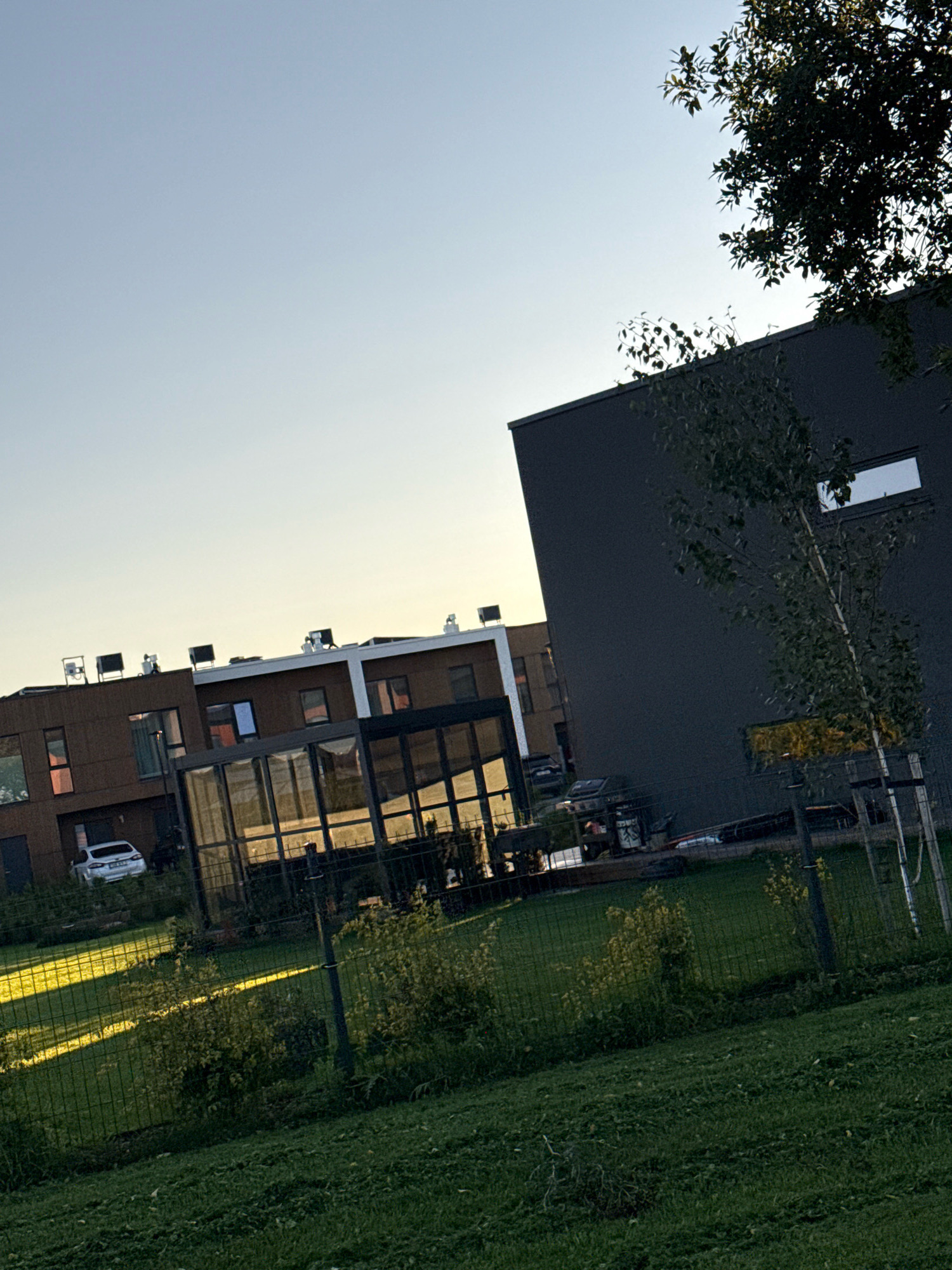 The image depicts a modern residential area in Järveküla, Estonia, during what appears to be late afternoon or early evening. The sky is a clear, pale blue with a hint of yellow near the horizon, suggesting the sun is low. In the foreground, a neatly mown green lawn slopes upwards towards a wire fence. Beyond the fence, a strip of lawn is illuminated by a bright ray of sunlight, casting a distinct diagonal shadow. Behind this sunlit area, there are several bushes with yellowing foliage.
Dominating the mid-ground are two contemporary buildings. The building on the left is clad in a warm, reddish-brown material and features multiple windows, some reflecting the sky. A white car is parked in front of this building. Adjacent to this structure is a glass-enclosed extension or sunroom, which reflects the surrounding environment, including the sunlight on the lawn.
To the right, a larger, dark gray building rises higher. This building has a minimalist design with a few horizontal, rectangular windows. A young, slender tree with sparse foliage stands in front of this dark building. The rooflines of the buildings are visible, some equipped with air conditioning units or similar rooftop equipment. The overall impression is one of modern architecture set within a suburban or semi-rural environment, characterized by clear lines, natural light, and green spaces.