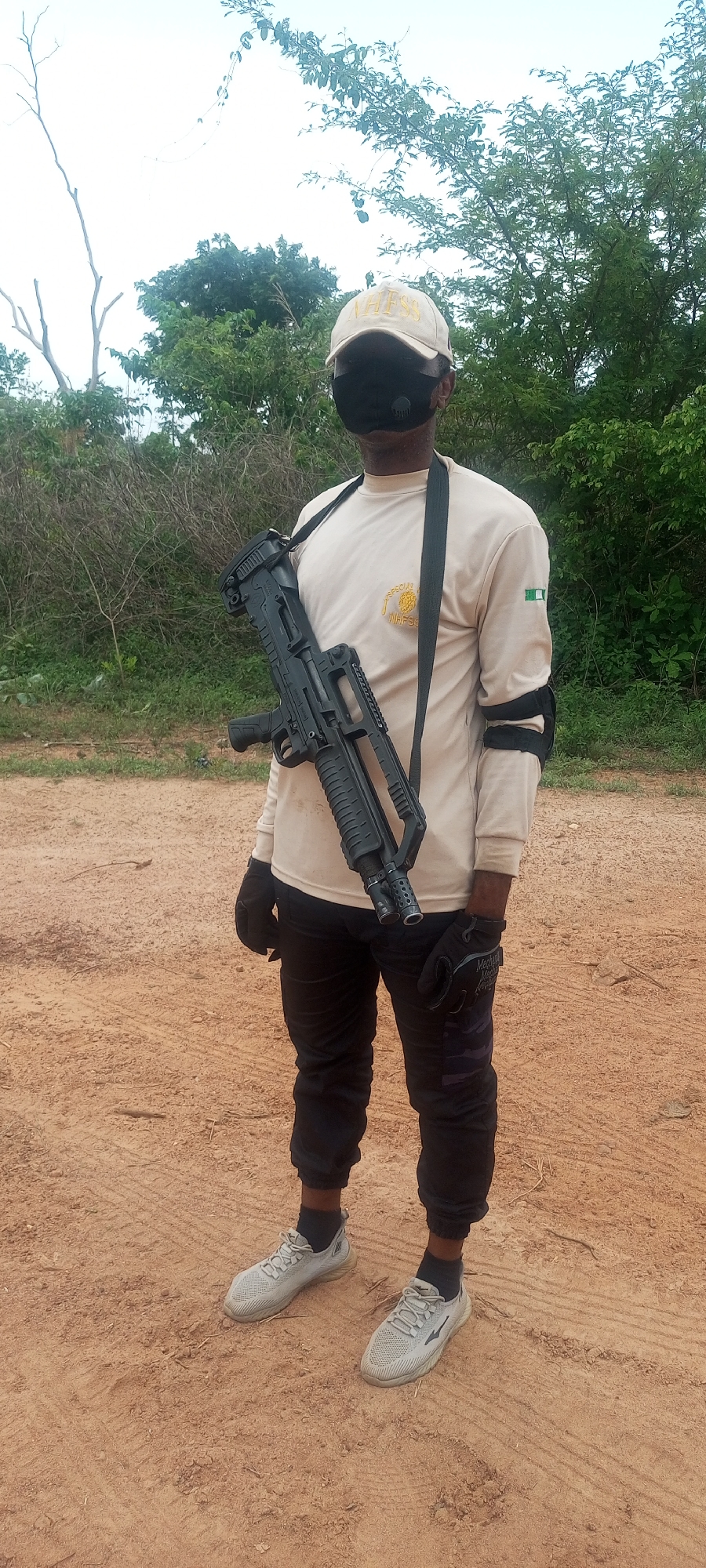 This full-length, eye-level portrait depicts a person standing outdoors on a dirt path in Otunade, Nigeria. The individual, a dark-skinned male, is dressed in what appears to be a uniform or tactical gear. He wears a light-colored (khaki or tan) long-sleeved shirt, black pants, black gloves, and grey athletic shoes. A beige baseball cap with the word "ATHESS" embroidered on the front covers his head, and a black face mask with a visible filter valve obscures his lower face.
A black bullpup-style shotgun or tactical rifle, characterized by its twin muzzle ports, is slung across his chest with a black strap over his left shoulder. On the left chest of his shirt, there is a circular gold emblem with the text "SPECIAL NHFSCS". The left sleeve displays a patch of the Nigerian flag. Black armbands or elbow guards are visible on both arms, and the right glove clearly shows the brand "Mechanix Wear."
The setting is an outdoor, rural or semi-rural environment with a reddish-brown dirt ground. Sparse green trees and bushes are visible in the background, beneath a bright, likely overcast sky, indicating daytime. The person stands in a static pose, appearing to be photographed. The overall impression is one of a security operative or someone engaged in a tactical exercise.