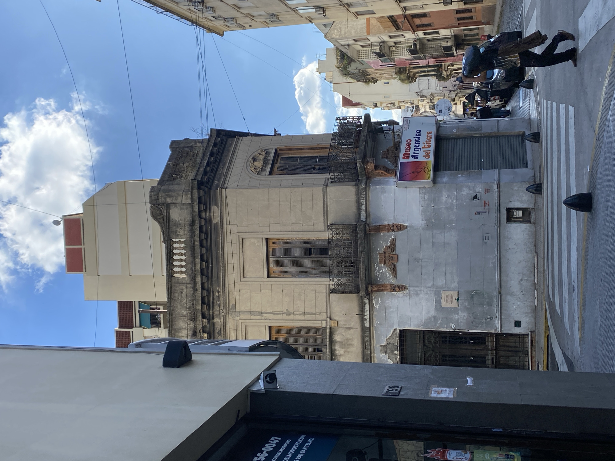 A street scene in Buenos Aires, Argentina. A narrow street view shows buildings on either side. On the right, a building with a partially damaged facade is visible; it has a closed roll-up door with the signage "Museo Argentino del Titere" (Argentine Puppet Museum) above it. A person in dark clothing is walking across a zebra crossing. The street is framed by buildings on both sides, with utility lines visible overhead against a blue sky with clouds. The building on the left has a sign with text in Spanish. A number "98" is visible on the front of the building.