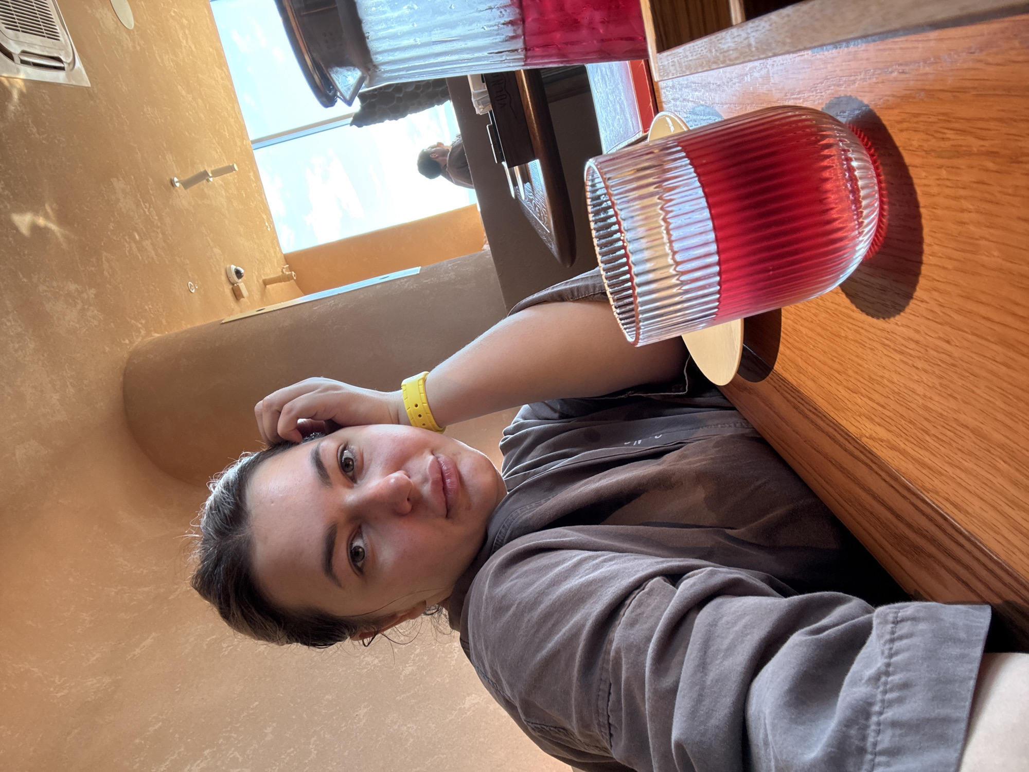 A young woman with her hand on her head and wearing a yellow bracelet poses for a selfie in a restaurant. She is sitting at a wooden table with a glass of red drink and a pitcher of red drink. The restaurant has a warm, earthy tone in its decor, with curved walls and a ceiling with exposed fixtures. Large windows allow natural light to enter, revealing a view of the sky. Another person can be seen in the background, seated at a table. The lighting suggests it is daytime.