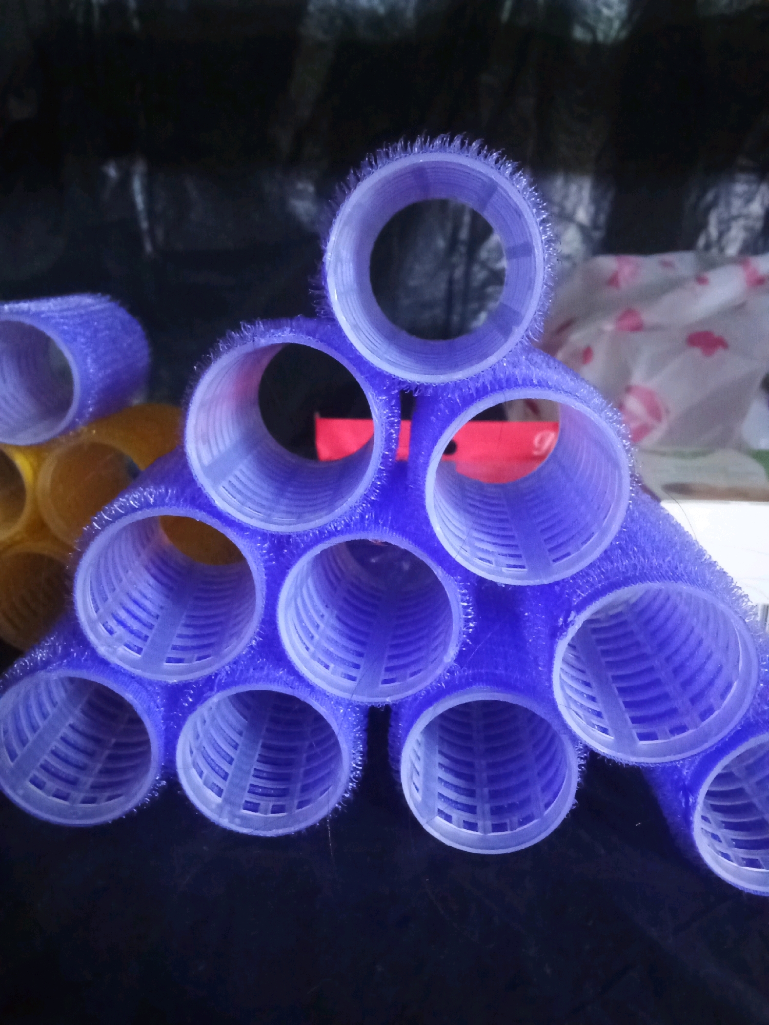The image displays a close-up, overhead view of several hair rollers. The rollers are predominantly purple, with some yellow rollers visible in the background. They are arranged in a triangular formation, with the purple rollers forming the apex of the triangle. Each roller is cylindrical and appears to be made of a textured material that would grip hair. The background is dark and indistinct, with some lighter, patterned material also visible. The lighting suggests an indoor setting, possibly a bathroom or vanity area. There are no people or discernible actions taking place. The image does not provide any information that could link it to Tbilisi, Georgia, or any specific city.