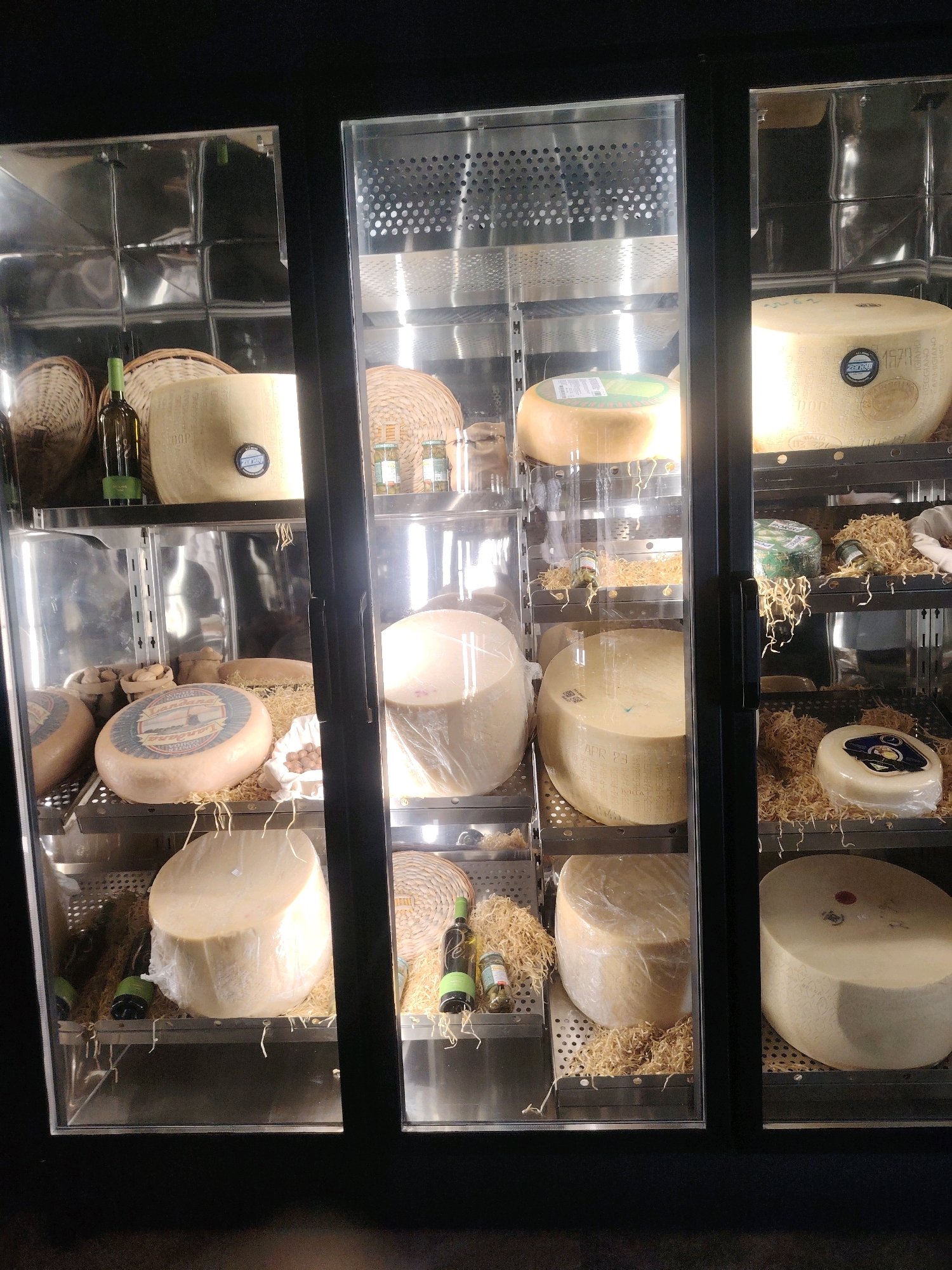 The image shows a refrigerated display case filled with various types of cheese. The display case is divided into three vertical sections, each with multiple shelves. The shelves are made of perforated metal, and some of the cheese wheels are placed on a bed of shredded straw-like material. Several large, round cheese wheels are prominently featured, some wrapped in plastic. Smaller cheeses, bottles of wine, and jars are also visible on the shelves. The lighting is bright, illuminating the cheeses and the metallic interior of the display case. The background is dark, suggesting an indoor commercial setting. There are no people visible in the image. The location is identified as Konigil, Uzbekistan.